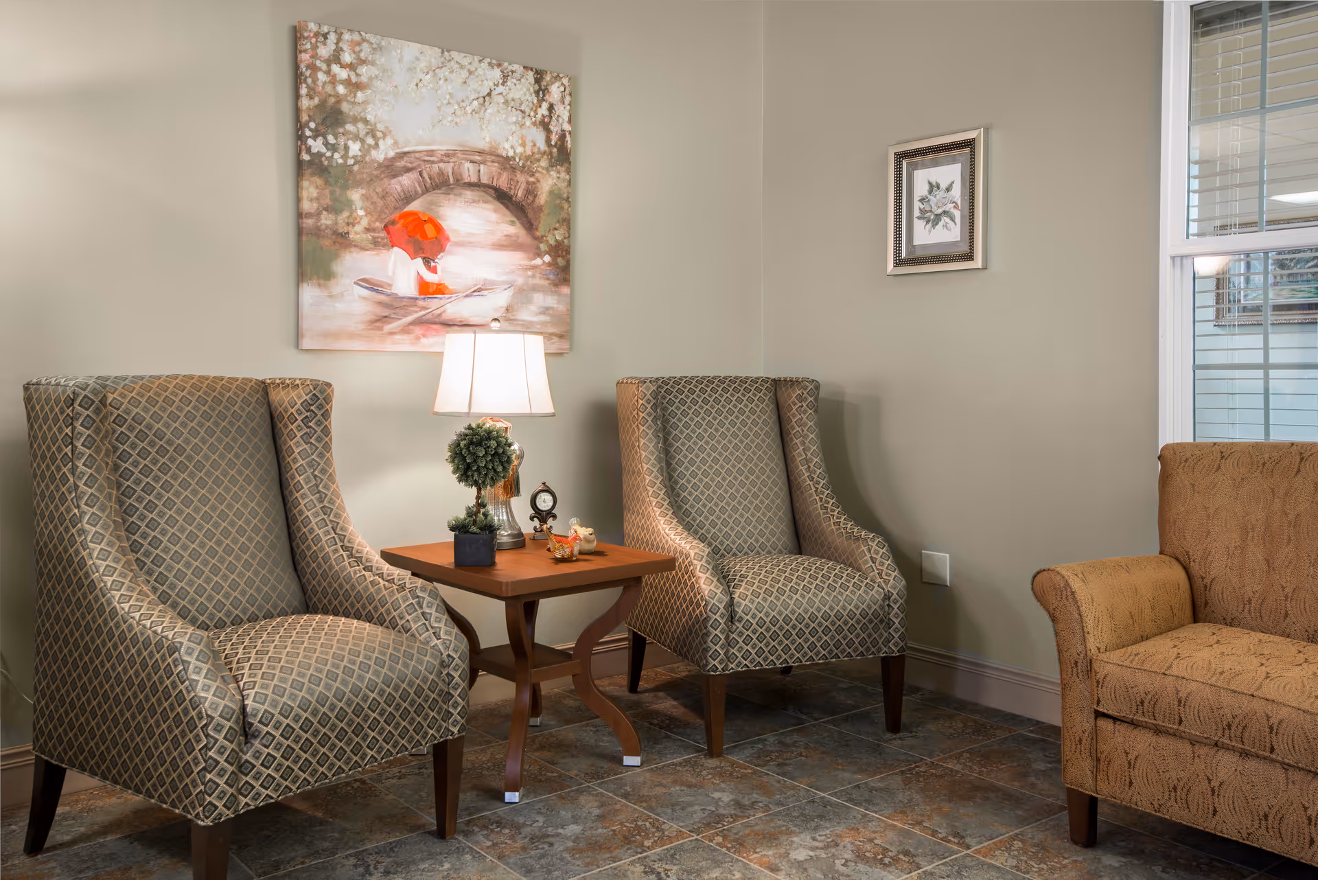 Two patterned armchairs flanking a wooden side table with a lamp and decorations in a cozy sitting area with wall art and a window.