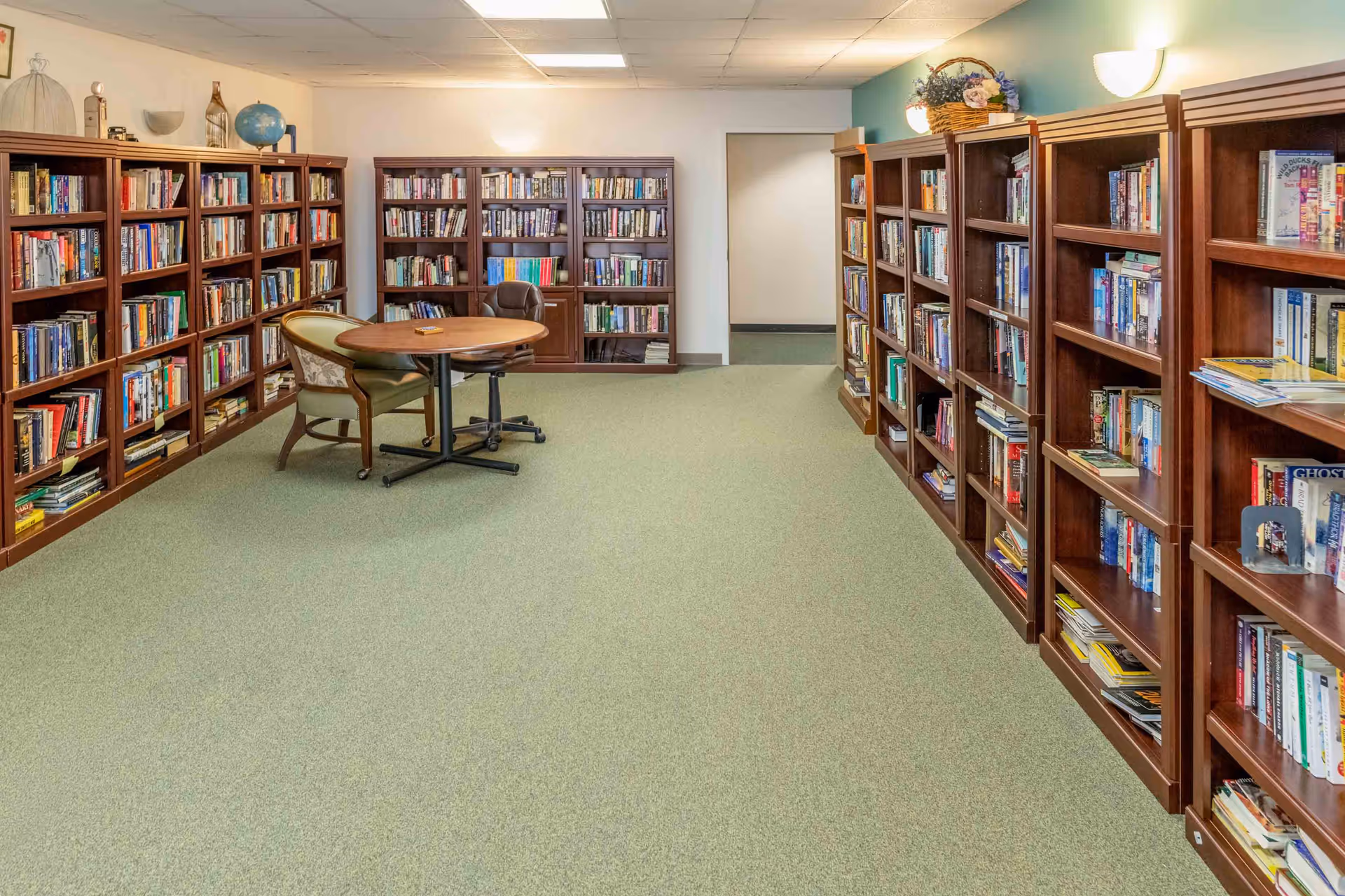 A quiet library room with wooden bookshelves lining the walls and a round table with chairs in the center.