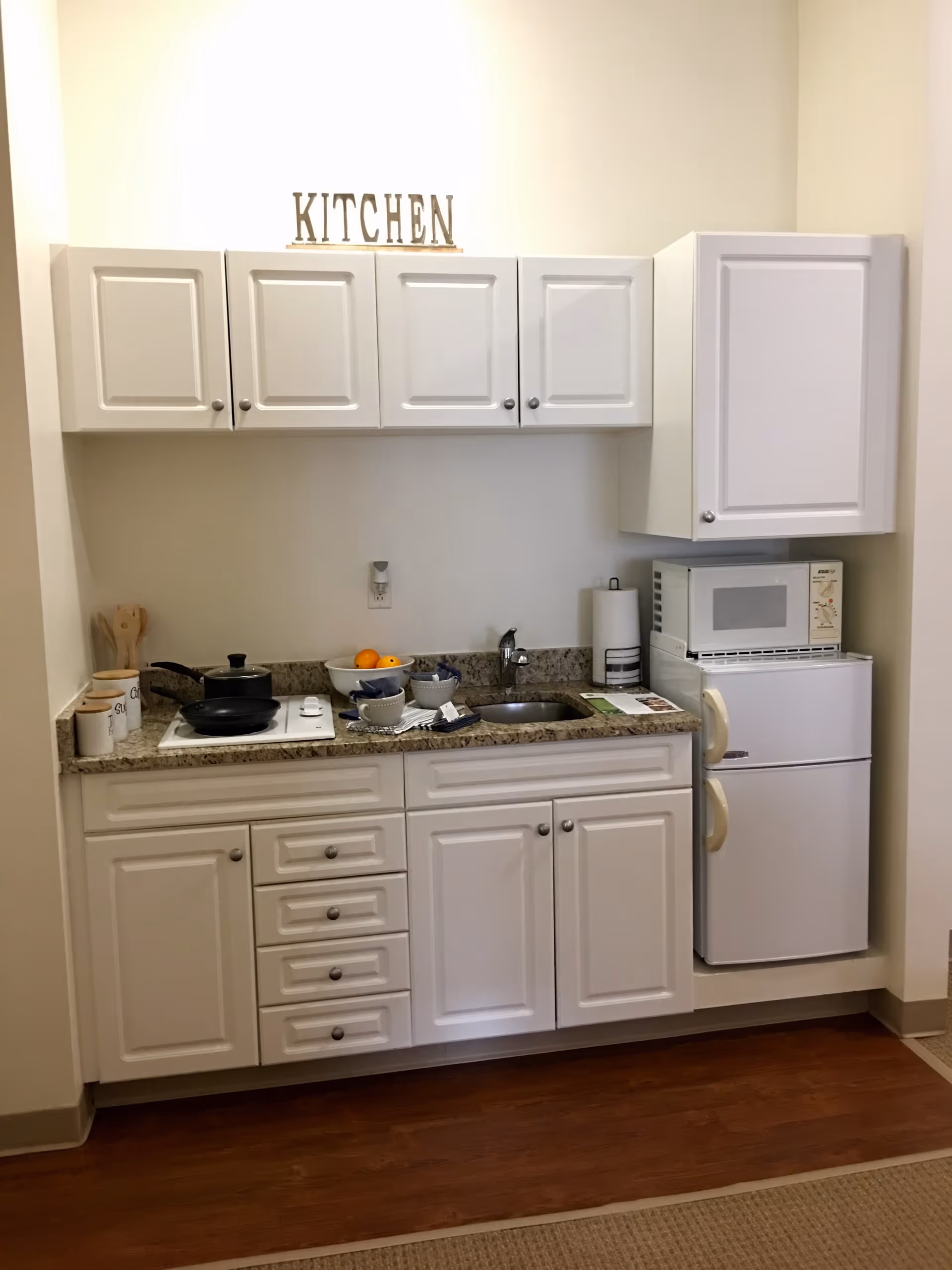 A small kitchen area with white cabinets and drawers, a granite countertop, a sink, a microwave on top of a mini refrigerator, and a stovetop with a frying pan and pot. There are kitchen utensils in a container, a bowl of fruit, and some cups on the counter. Above the cabinets, there is a decorative sign that reads 'KITCHEN'.