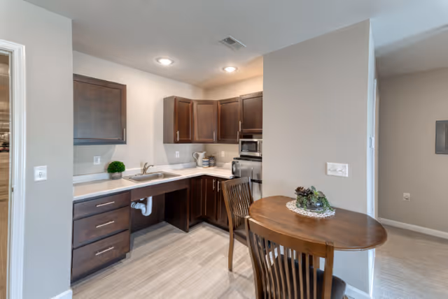 A small kitchen area with dark wood cabinets, a stainless steel microwave and oven, a sink, and a small round wooden table with two chairs. The kitchen has light-colored countertops and flooring, with neutral-colored walls.