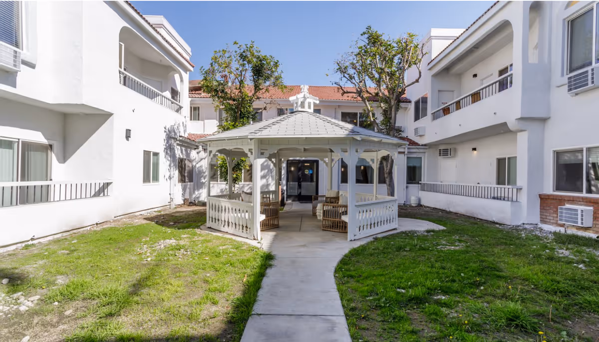 A white gazebo sits in the center of a grassy courtyard surrounded by a two-story white senior living building.