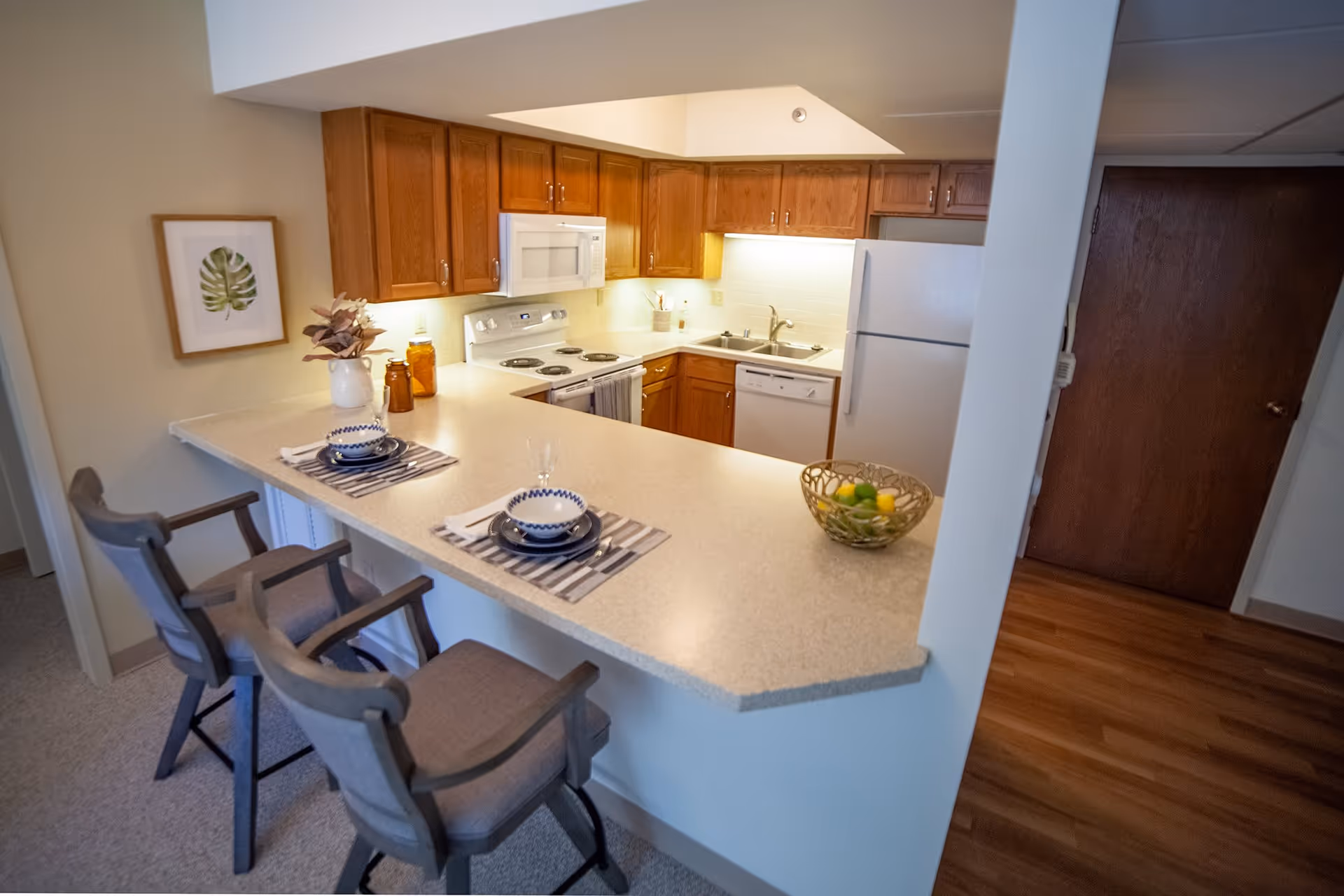 A small kitchen area with wooden cabinets, a white refrigerator, stove, microwave, and dishwasher. A countertop extends outward with two place settings, each with a bowl, plate, and glass, and two cushioned chairs. A framed leaf artwork hangs on the wall, and a bowl of fruit is on the counter.