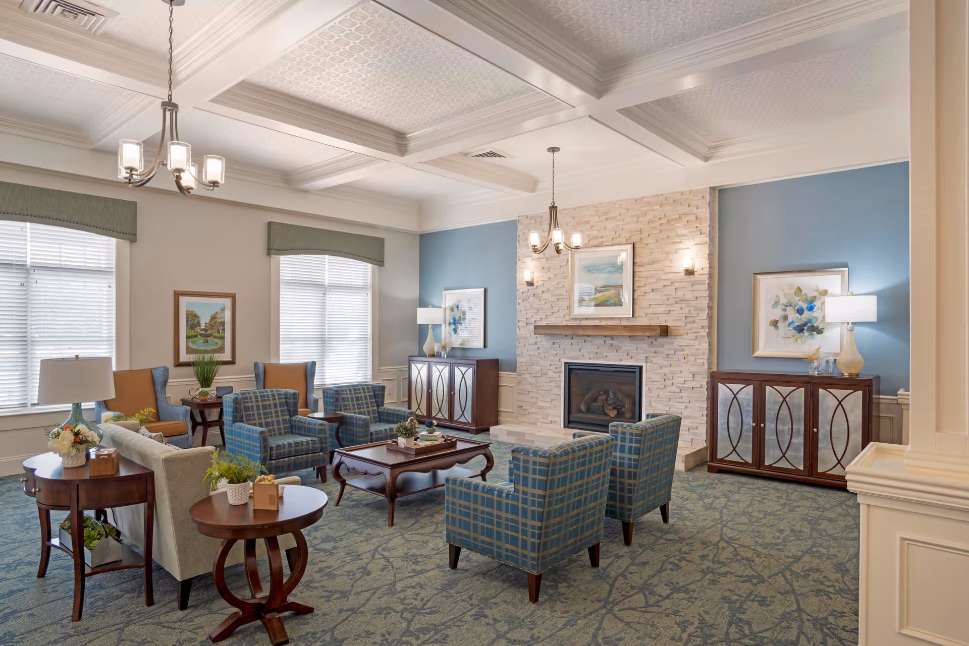 A cozy living room with a stone fireplace centered on a blue accent wall. The room features a mix of seating including a beige sofa, four blue plaid armchairs, and two brown wingback chairs. There are wooden side tables with lamps and plants, framed artwork on the walls, and two chandeliers hanging from a coffered ceiling. The carpet has a subtle floral pattern.