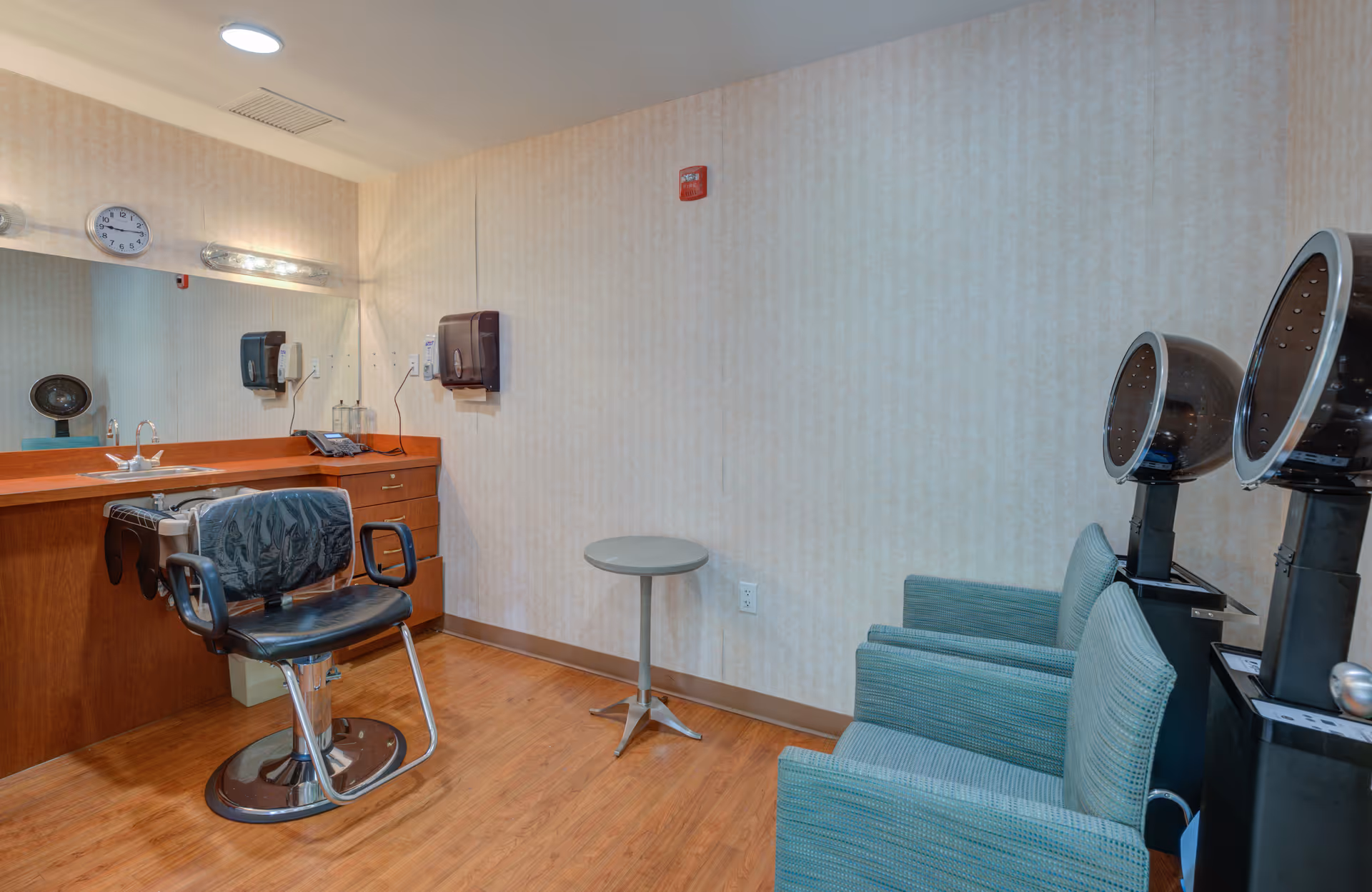 Interior view of a salon area in a senior living facility featuring a black salon chair in front of a wooden counter with a large mirror, a clock on the wall, and two hair dryers with green upholstered chairs beside them. The room has wood flooring and beige patterned wallpaper.