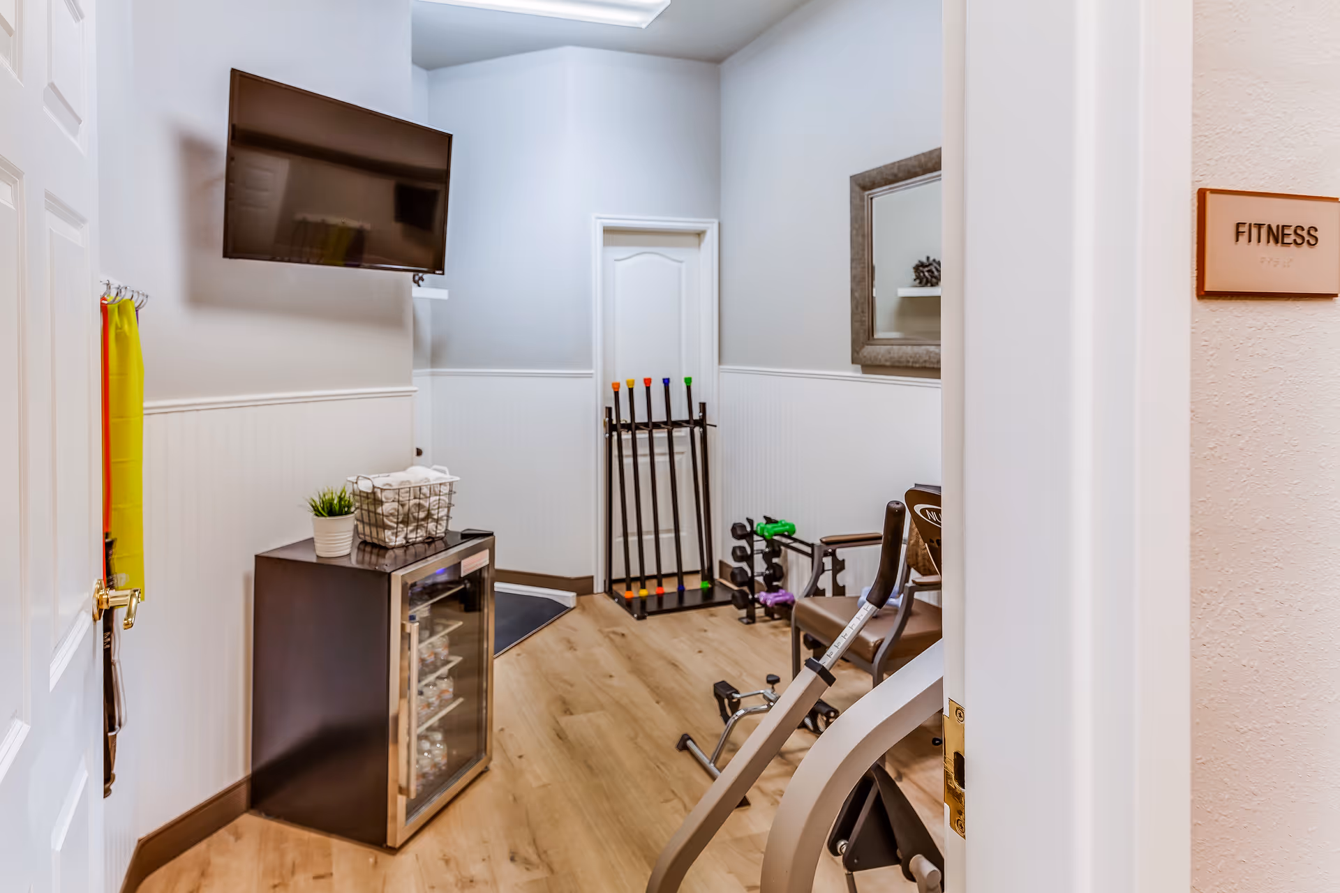 Small fitness room with exercise equipment including a stationary bike, dumbbells, and resistance bars. A flat-screen TV is mounted on the wall above a small refrigerator stocked with water bottles. A mirror hangs on the wall and a sign on the door reads 'FITNESS'.