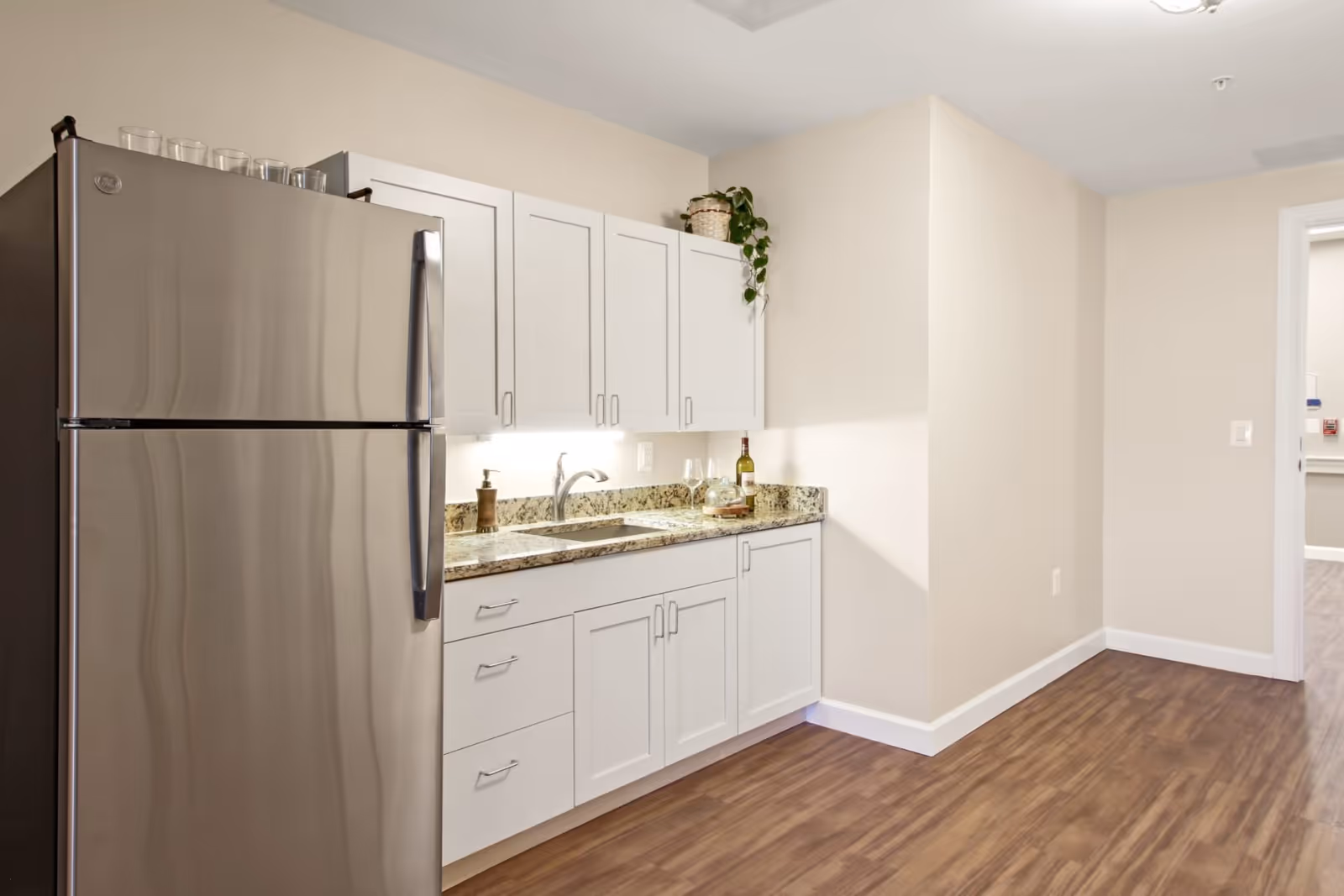 Small kitchen area with stainless steel refrigerator, white cabinets, granite countertop and sink on wood-look flooring.