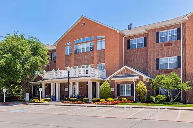 Exterior view of a three-story brick building with white trim and black shutters, featuring a covered entrance with white columns and a balcony above. The building is surrounded by landscaped greenery and colorful flowers, with a parking area in front under a clear blue sky.