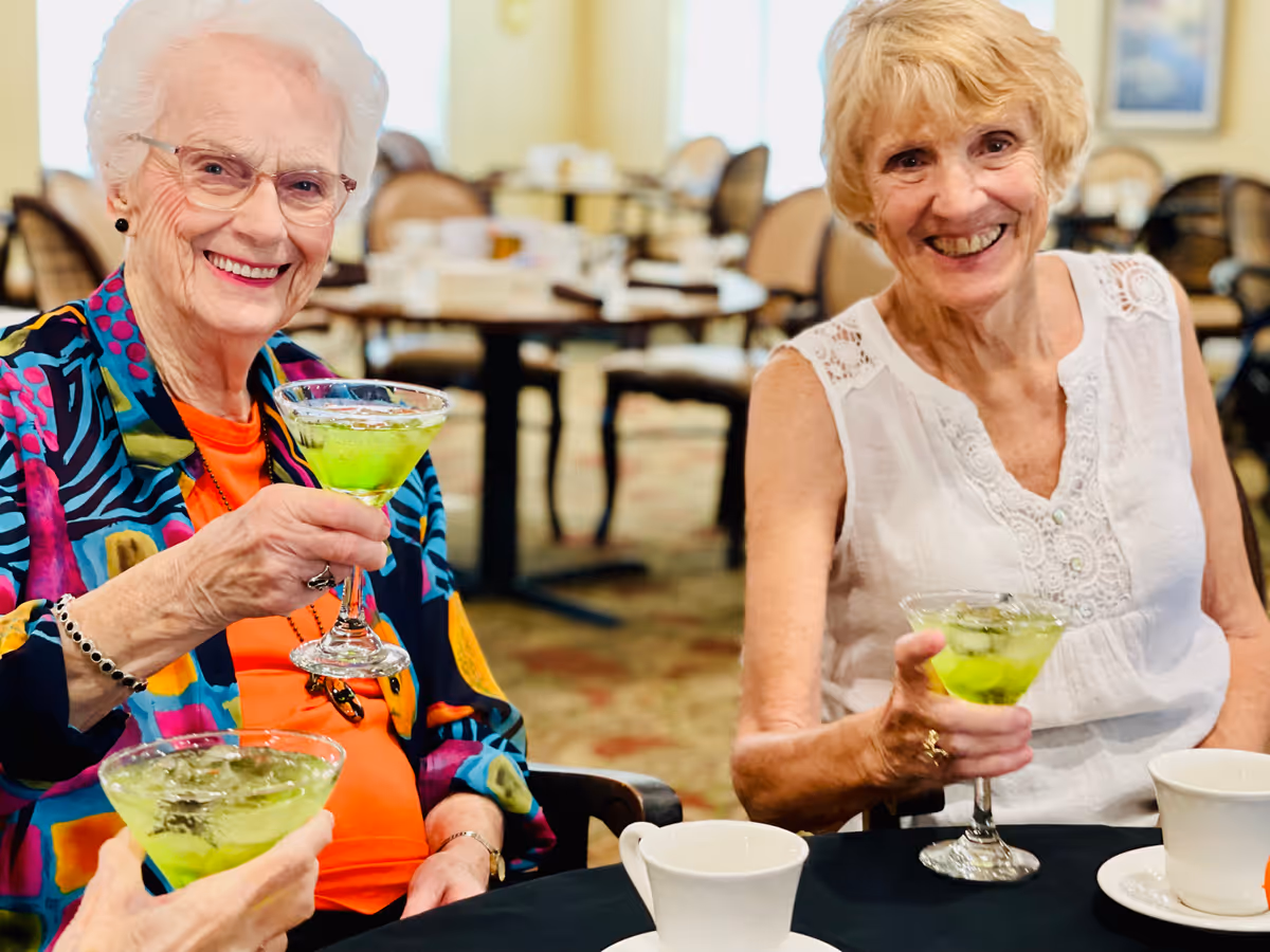 Two elderly women sitting at a table in a dining area, smiling and holding glasses with a green beverage. The background shows more tables and chairs in a well-lit room.
