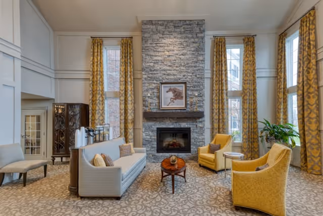 A spacious and elegant living room area with a tall stone fireplace centered on the back wall. The room features large windows with yellow patterned curtains, a beige sofa with decorative pillows, two yellow armchairs, a small round wooden coffee table, and a side table with a plant. The carpet has a subtle floral pattern, and the walls are painted in neutral tones.