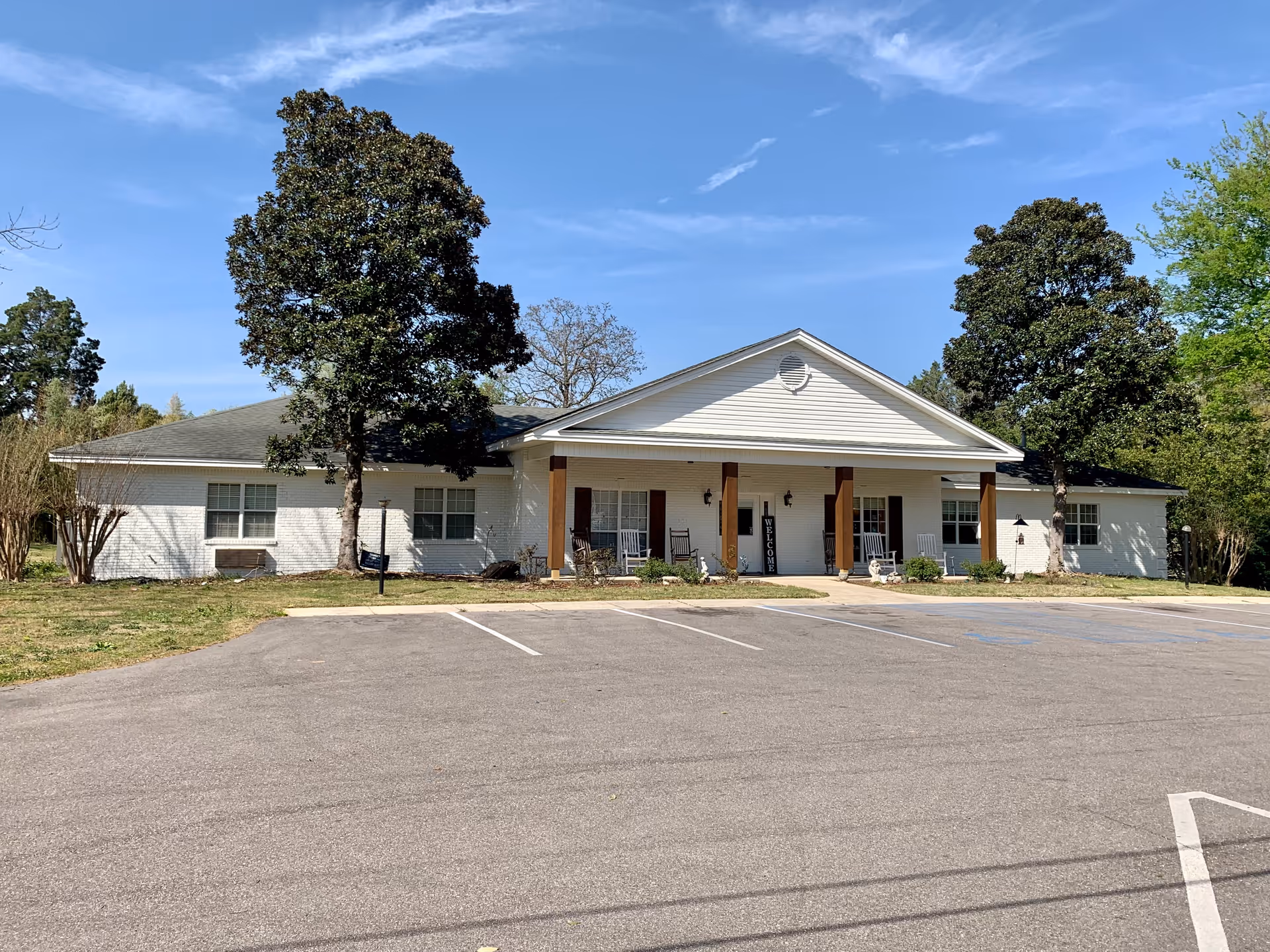 Front exterior view of a single-story white brick building with a covered porch supported by wooden columns. There are rocking chairs on the porch and a welcome sign near the entrance. The building is surrounded by trees and has a parking lot in front with marked parking spaces under a clear blue sky.