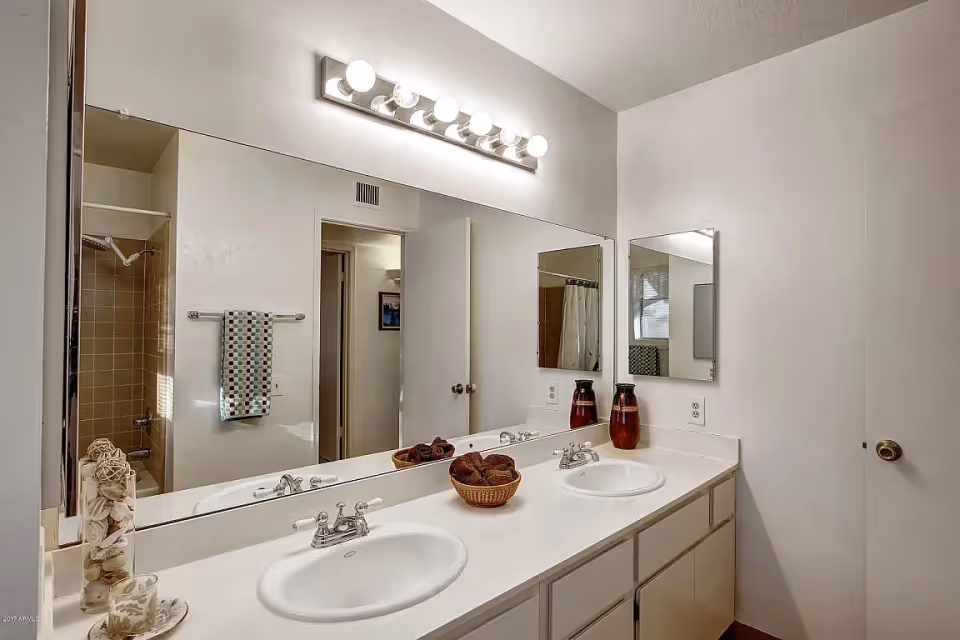 Double-sink bathroom vanity with a large mirror, overhead lights, decorative accents, and a towel near a shower entrance.