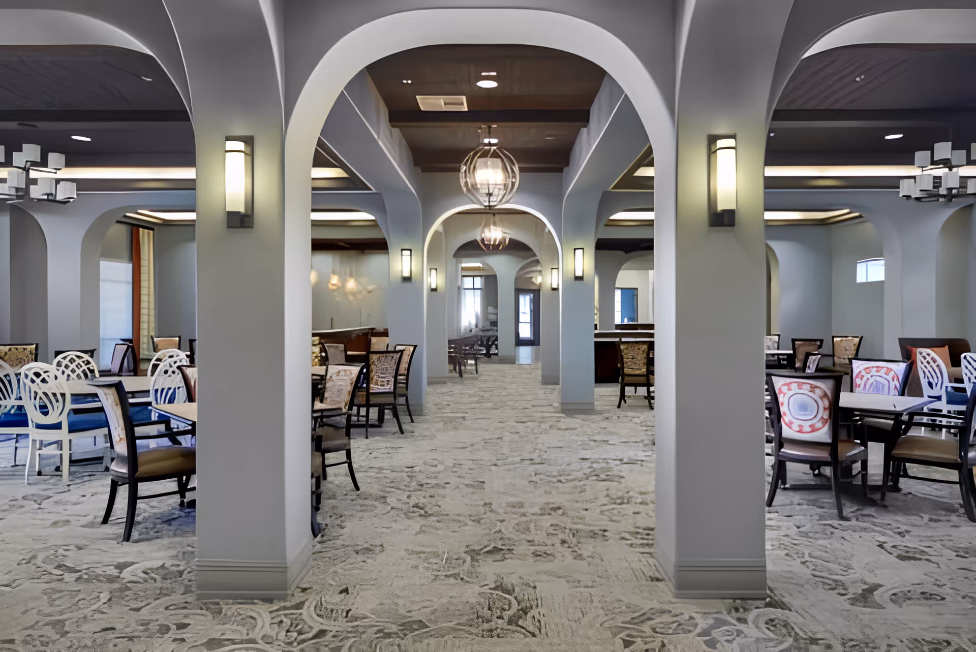 A spacious dining room with multiple tables and chairs arranged neatly. The room features arched columns, modern light fixtures on the walls and ceiling, and a patterned carpet floor. The overall color scheme is neutral with soft lighting.