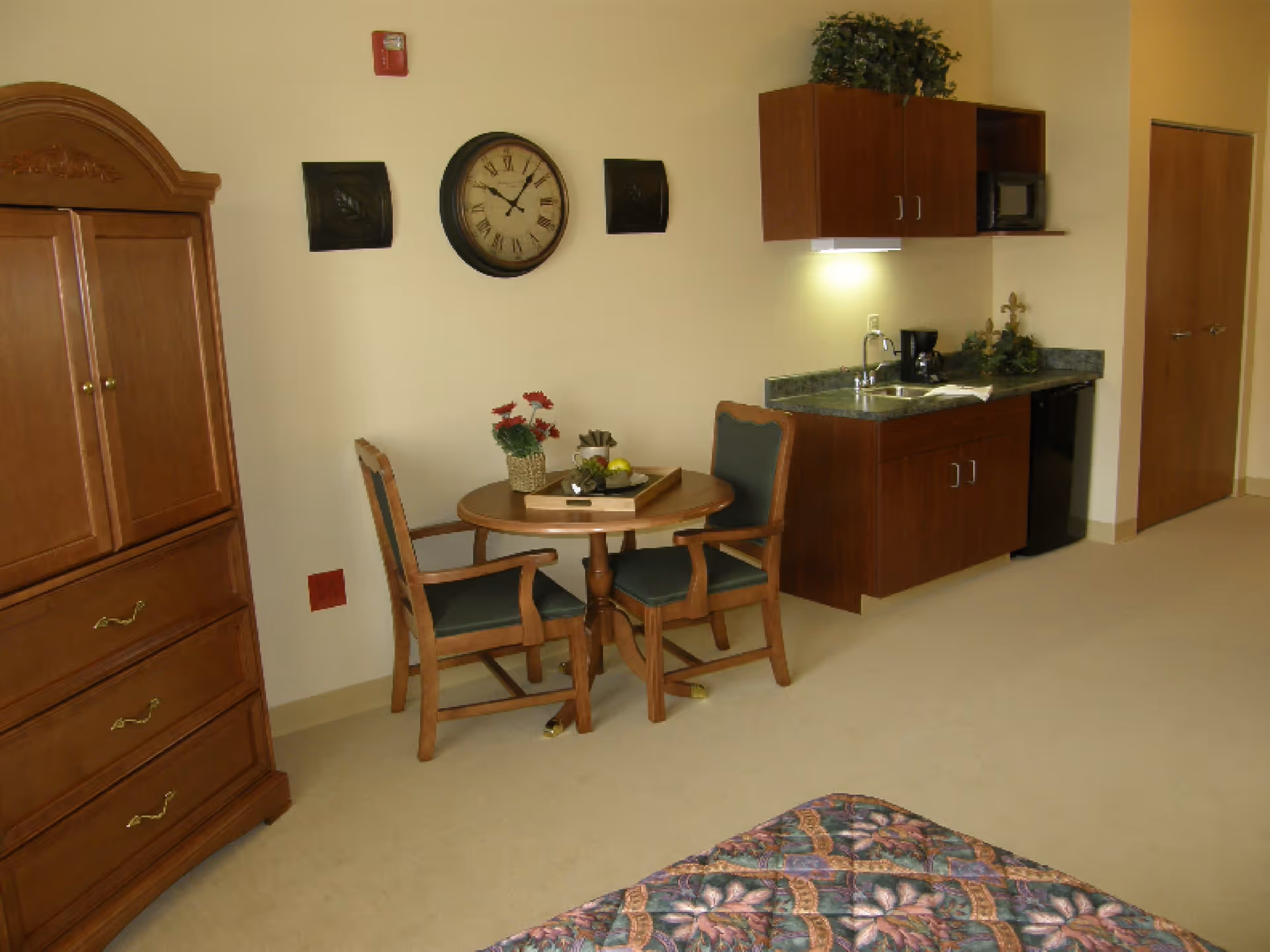 Studio apartment interior with a small kitchenette, round table and two chairs, wooden armoire, and part of a bed visible.