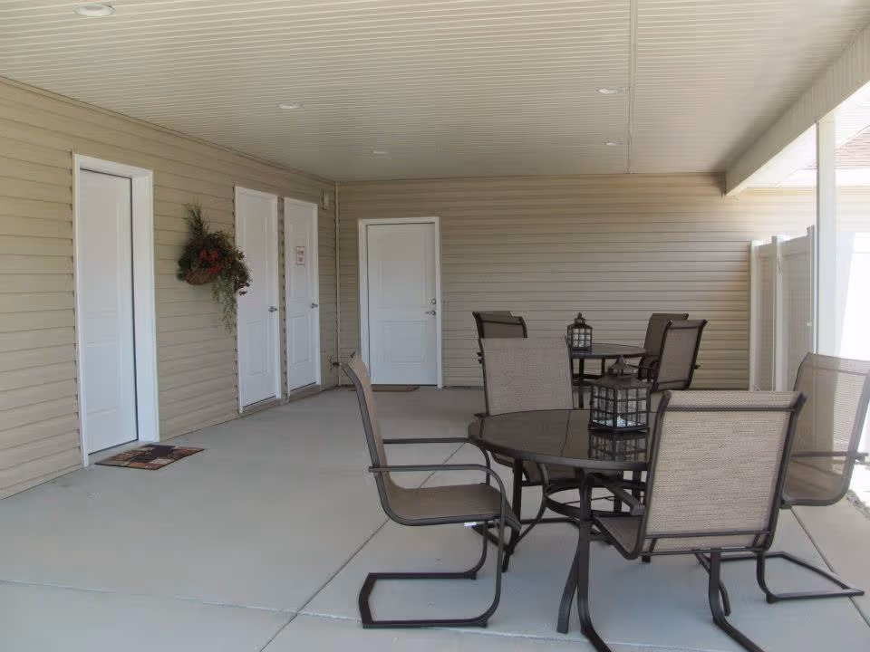 Covered outdoor patio area with beige siding walls, four white doors, and several round tables with brown mesh chairs. Each table has a decorative lantern on top. A hanging floral arrangement is mounted on the wall near the doors.