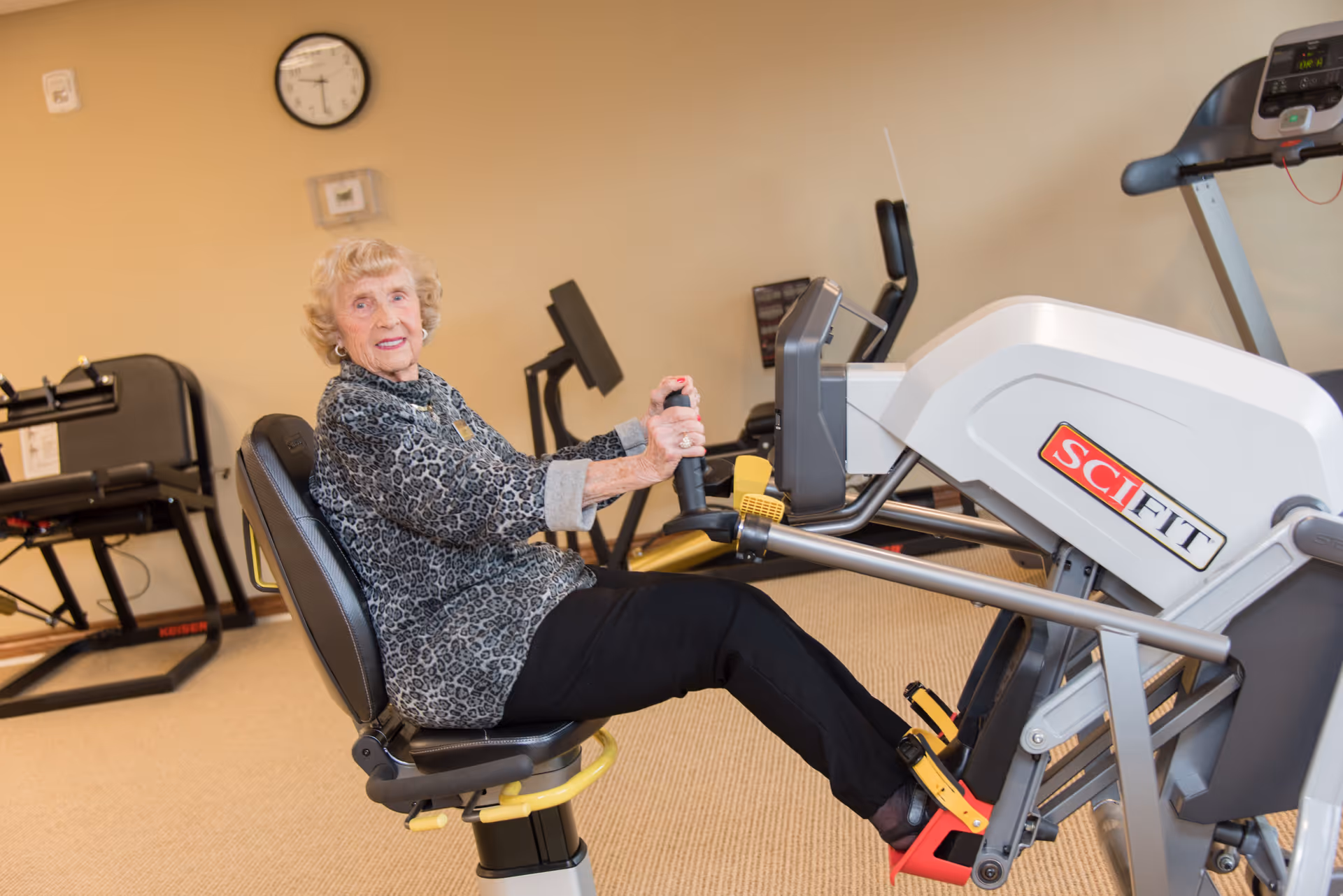 An elderly woman exercising on a seated rowing machine in a fitness room with other exercise equipment visible in the background.