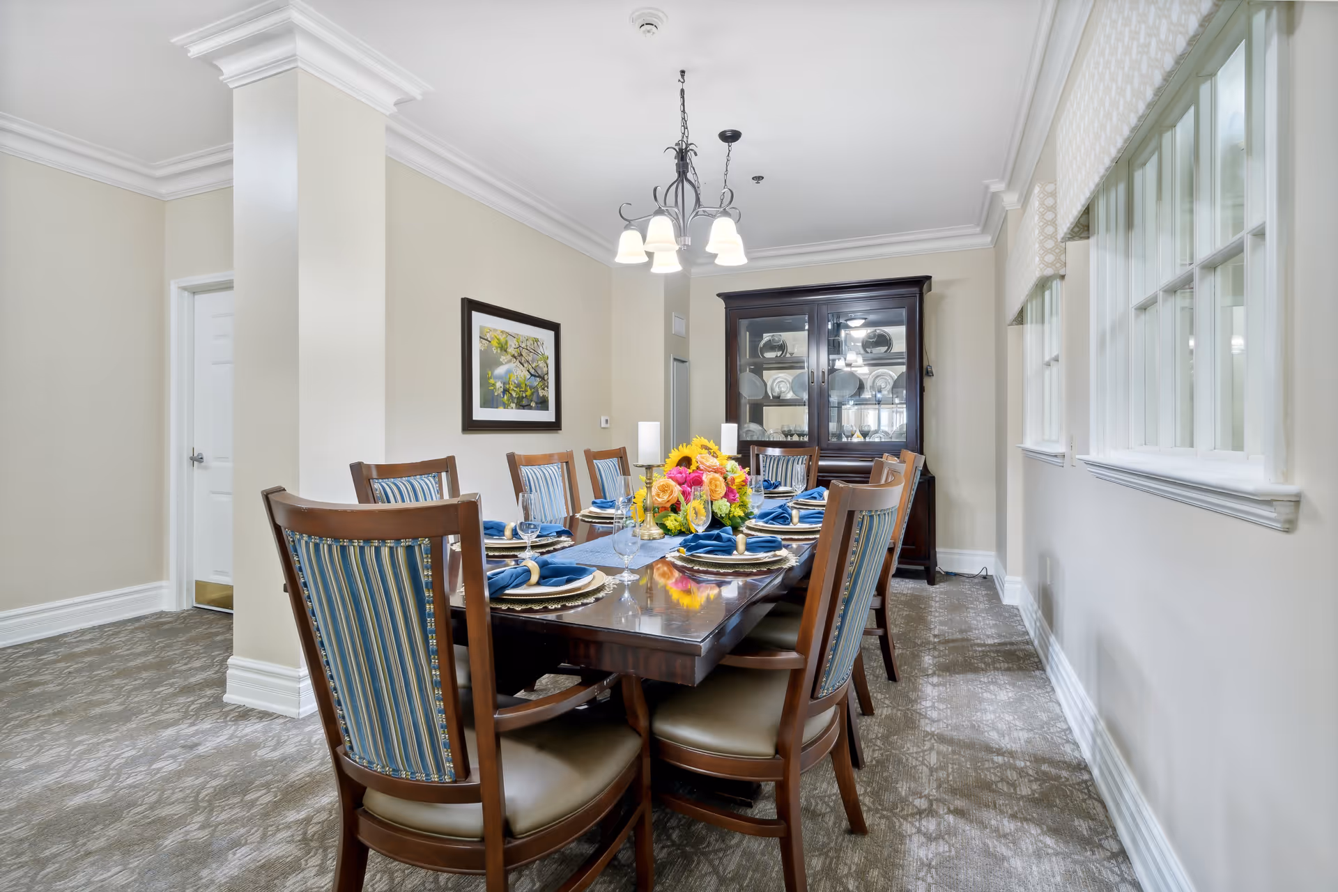 A formal dining room with a long wooden table set for eight people. The table is decorated with blue napkins, plates, and a centerpiece of colorful flowers. There are wooden chairs with blue striped upholstery around the table. A dark wooden china cabinet with glass doors displaying plates is against the far wall. The room has beige walls, a carpeted floor, and a chandelier hanging from the ceiling. Large windows with white trim and patterned valances are on the right side.