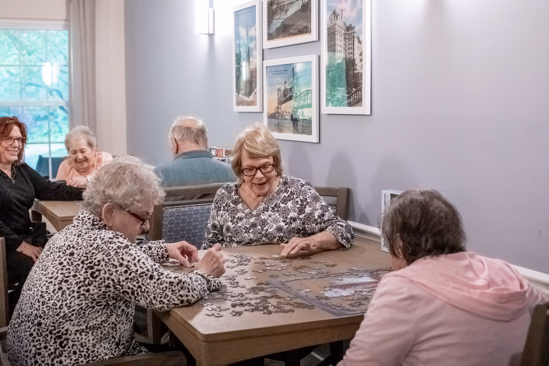 A group of elderly women sitting around a wooden table in a well-lit room, working together on assembling a jigsaw puzzle. The room has light gray walls decorated with framed pictures, and there is a window with curtains in the background.