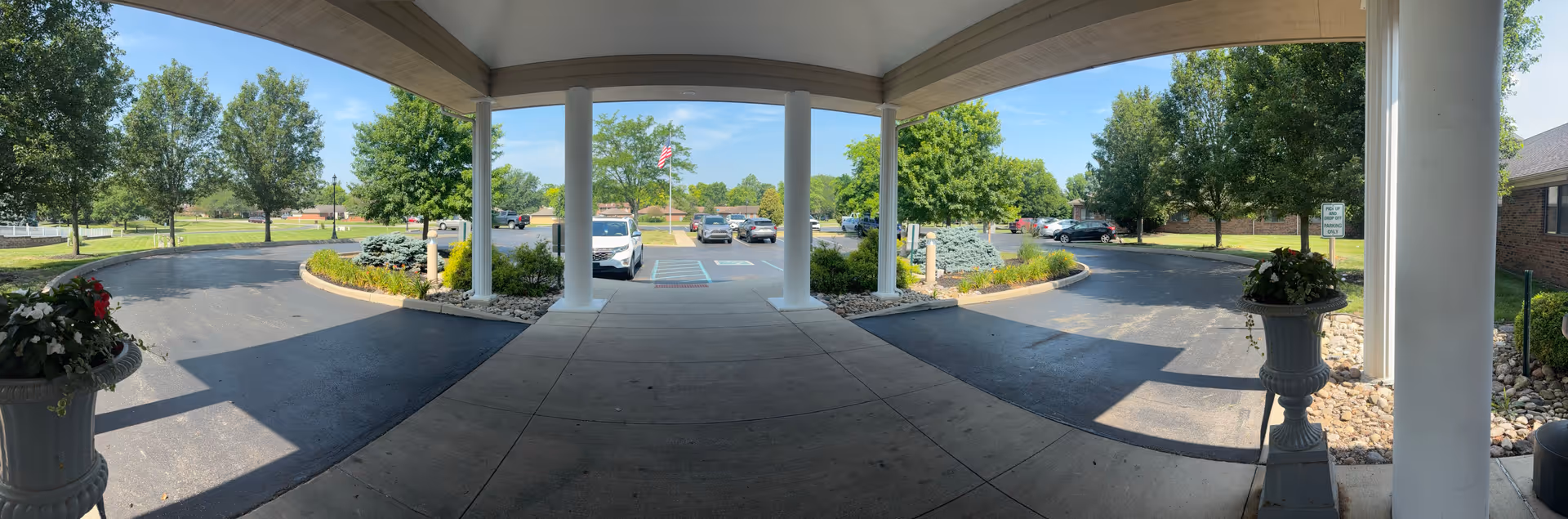 Covered entrance area of Walnut Creek Senior Living Campus with white columns, a circular driveway, landscaped greenery, and a parking lot with several cars. Trees and a clear blue sky are visible in the background.