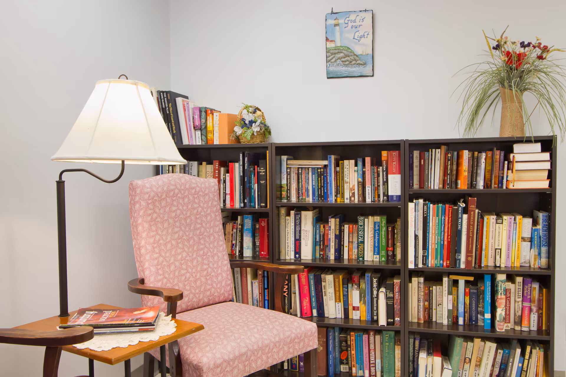 A cozy reading corner with a pink upholstered armchair next to a wooden side table holding magazines and a lamp. Behind the chair are three bookshelves filled with numerous books. A small floral arrangement is on top of one bookshelf, and a framed picture with the text 'God is our Light' hangs on the wall above.