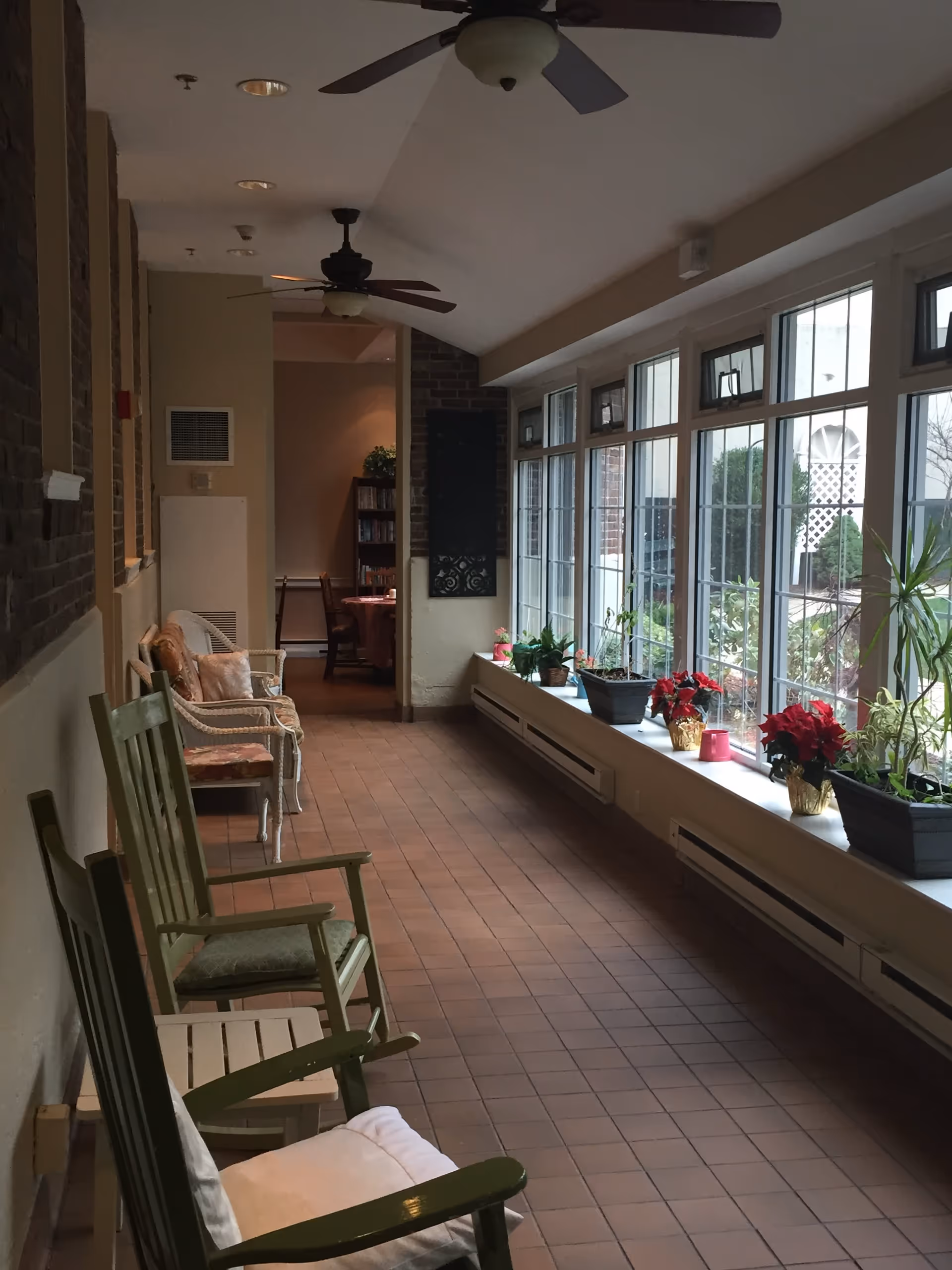 A long indoor hallway with large windows on the right side letting in natural light. The windowsill is decorated with various potted plants and flowers. On the left side, there are several chairs including green wooden rocking chairs and cushioned wicker chairs. Ceiling fans are mounted on the ceiling, and a small room with a table and chairs is visible at the end of the hallway.