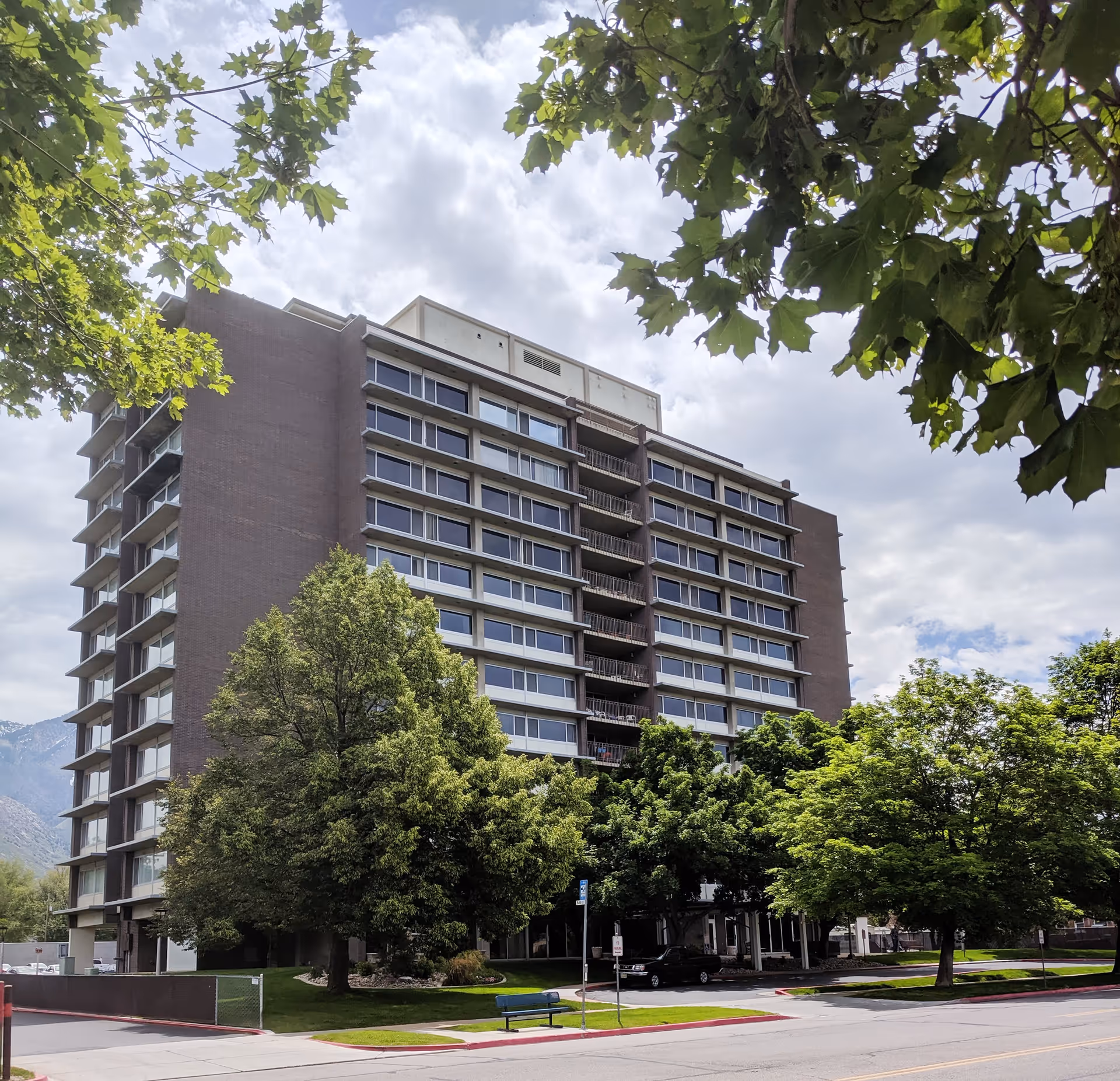 A multi-story residential building surrounded by green trees with a partly cloudy sky in the background. The building has large windows and balconies, and there is a street with a bench and parked cars in front.