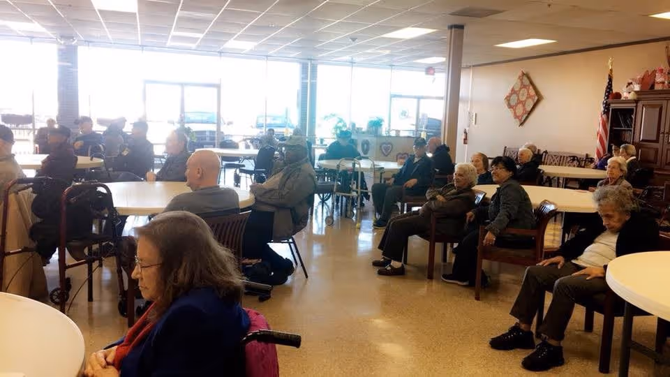 A group of elderly people seated around tables in a large, well-lit room with windows along one wall. Some individuals use walkers or wheelchairs. The room has a tiled ceiling, beige walls, and an American flag in the corner. The atmosphere appears calm and communal.