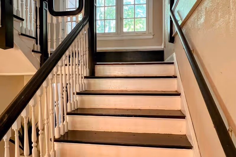 Wooden indoor staircase with dark treads, white risers and a spindled banister leading up to a window.