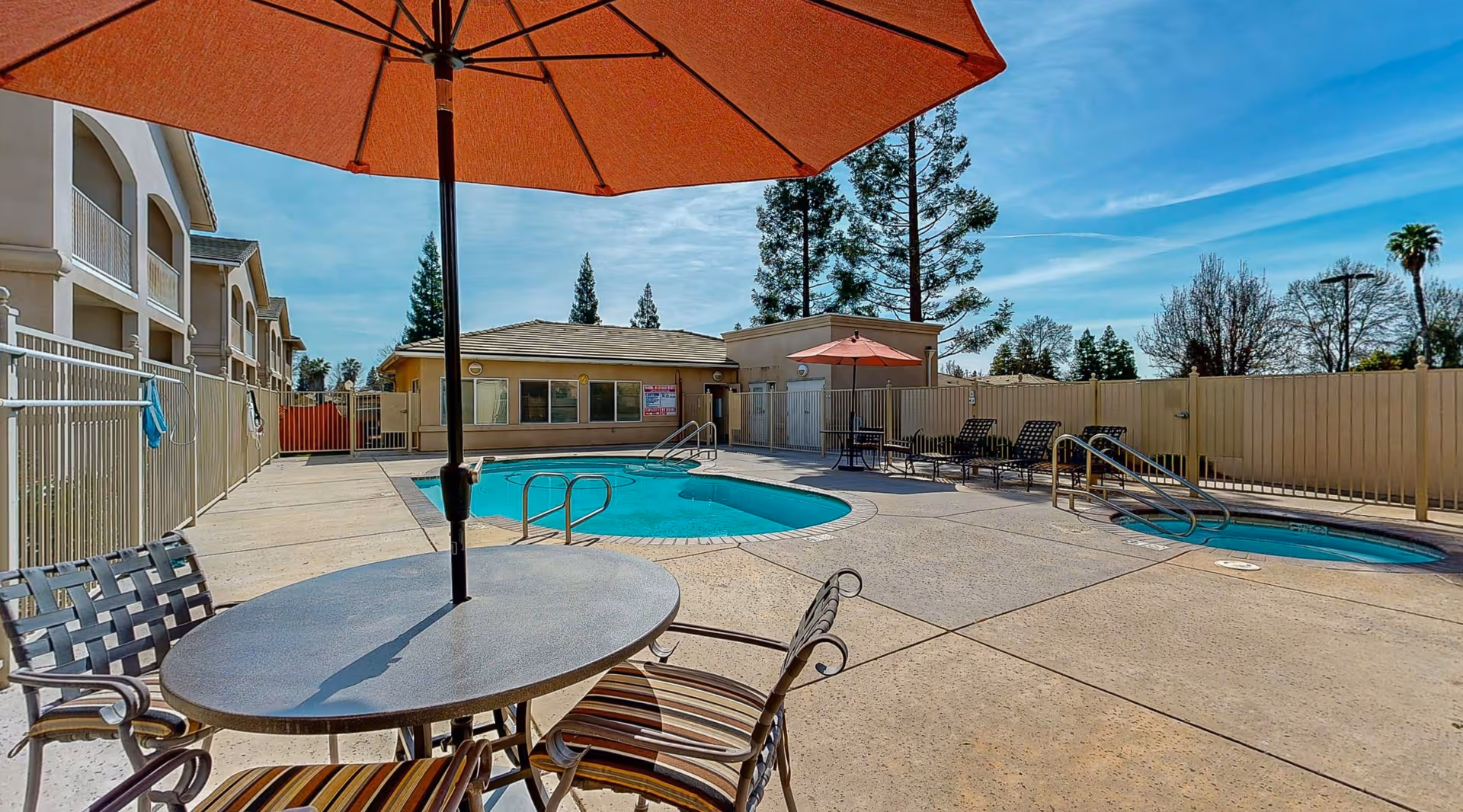 Outdoor pool area at Cogir of Cedar Creek with a round table and chairs under a large orange umbrella in the foreground. The pool is surrounded by a concrete deck with lounge chairs and another table with an umbrella. A beige building and a fence enclose the area, with trees and a blue sky in the background.