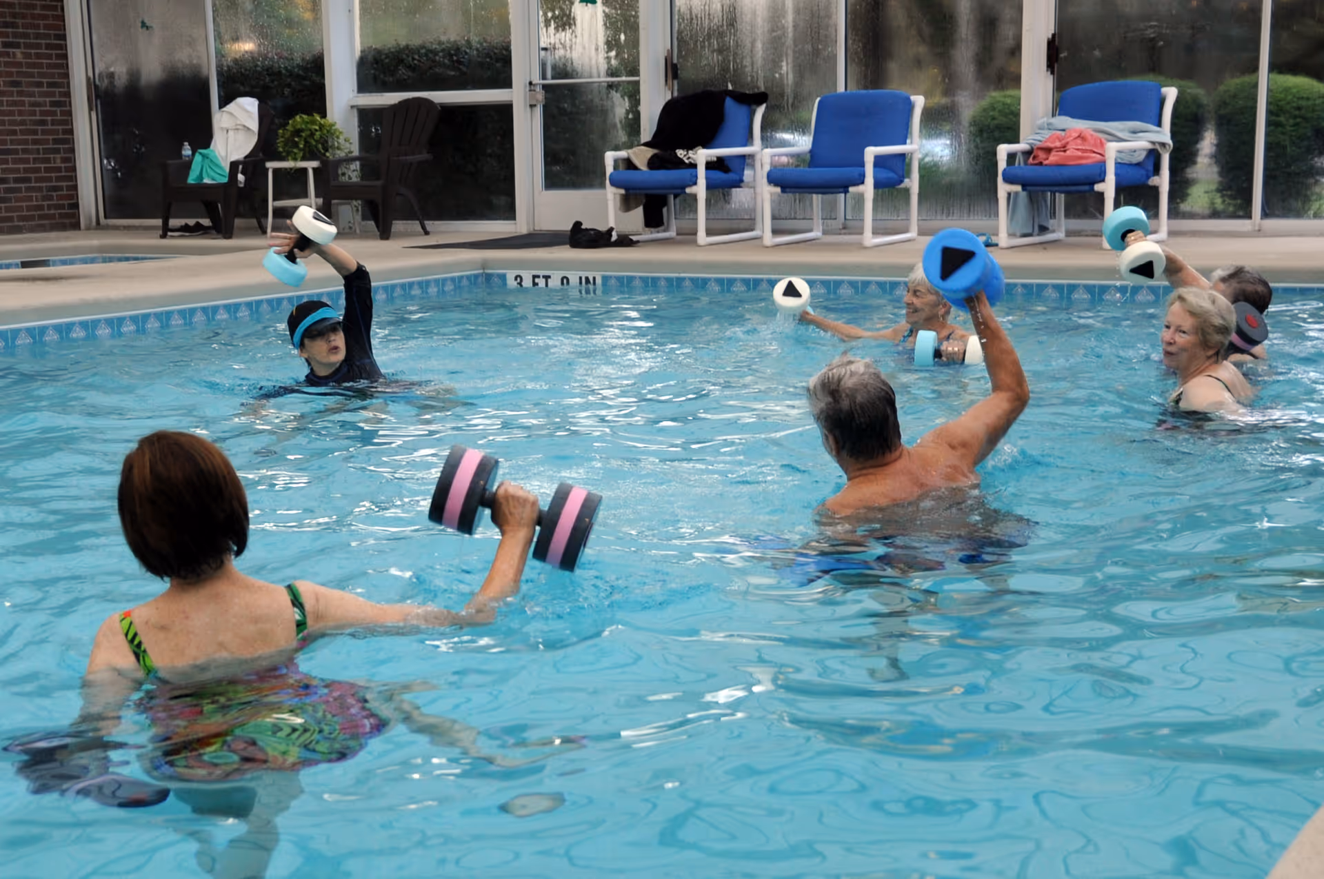 A group of elderly people participating in a water exercise class in an indoor swimming pool, each holding foam dumbbells. There are chairs and towels visible on the pool deck in the background.