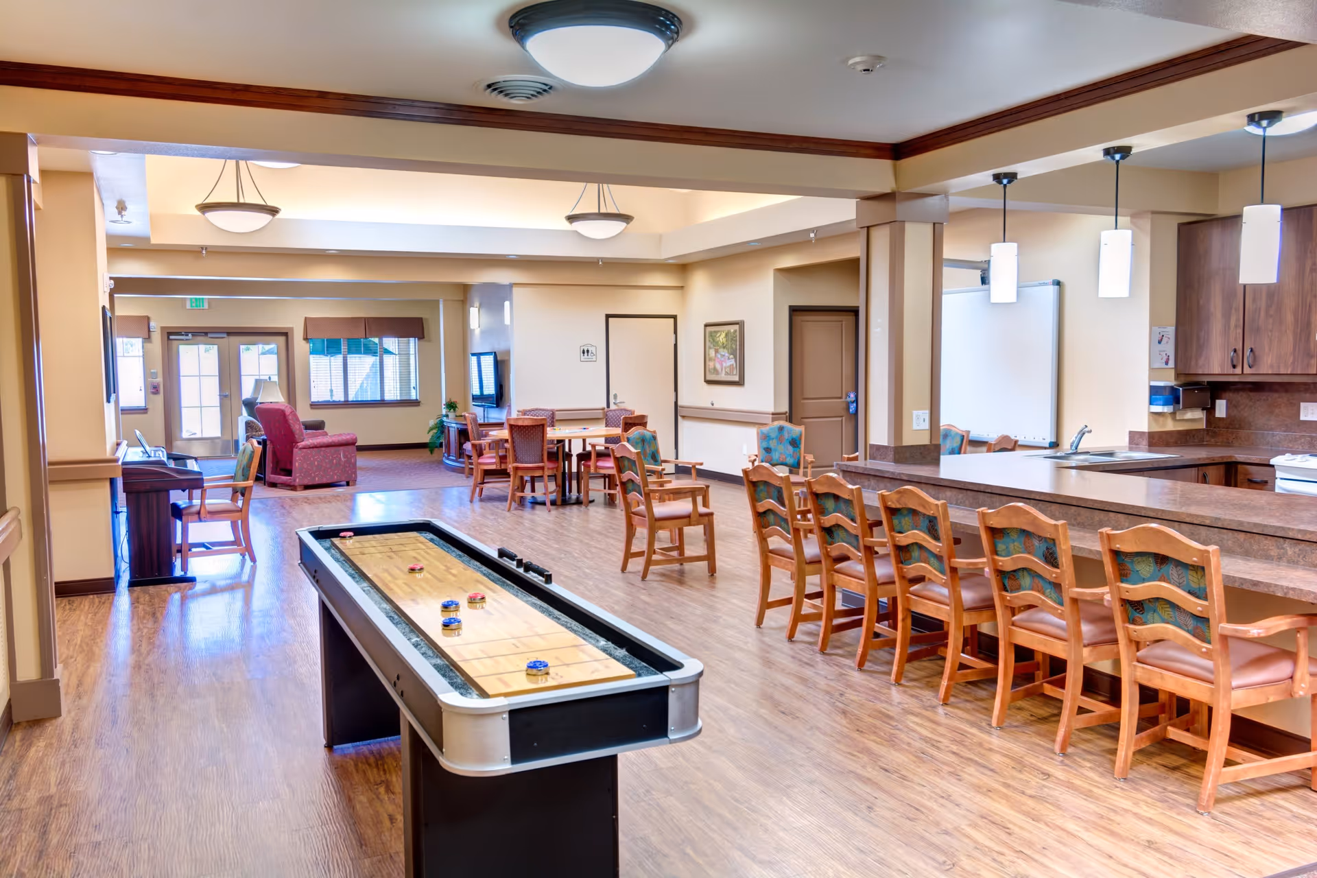 A spacious common area in a senior living facility featuring a shuffleboard table in the center, a long counter with wooden chairs on the right, and several tables and chairs in the background. The room has wood flooring, beige walls, and ceiling lights, with windows and a door letting in natural light.