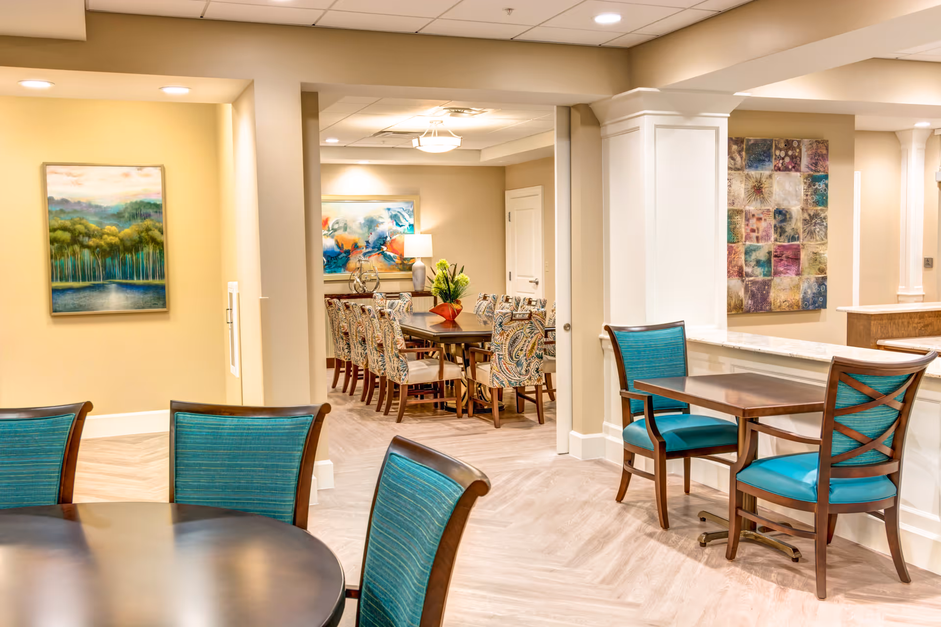 Interior view of a senior living facility dining area with multiple tables and chairs. The foreground shows a round table with teal cushioned chairs, and to the right, a small square table with two matching chairs. In the background, there is a larger dining table with patterned chairs and decorative artwork on the walls. The space is well-lit with ceiling lights and has a warm, inviting atmosphere.