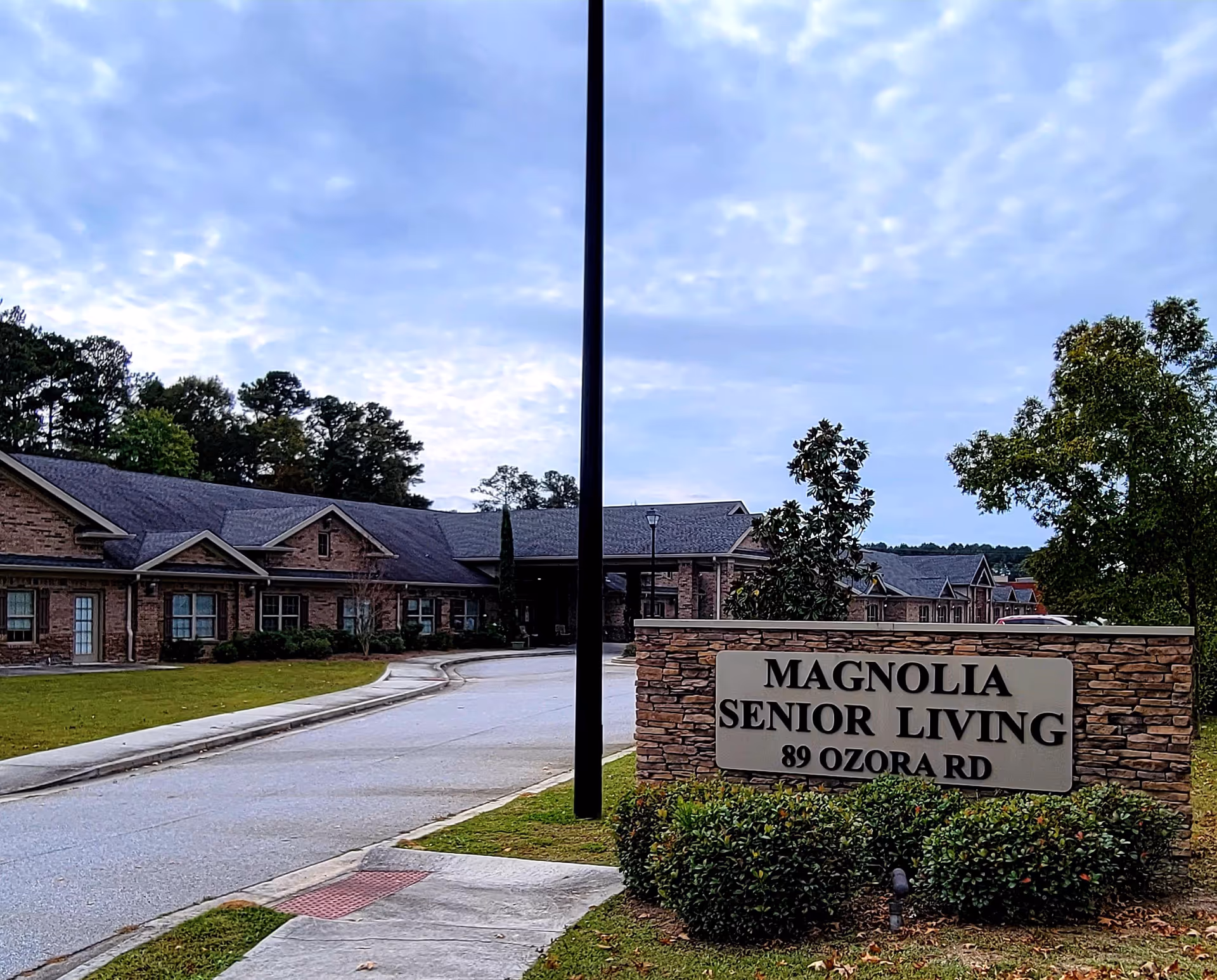 Exterior view of Magnolia Senior Living facility with a stone sign displaying the name and address 89 Ozora Rd. The building is a single-story structure with a brick facade, surrounded by trees and greenery under a cloudy sky.