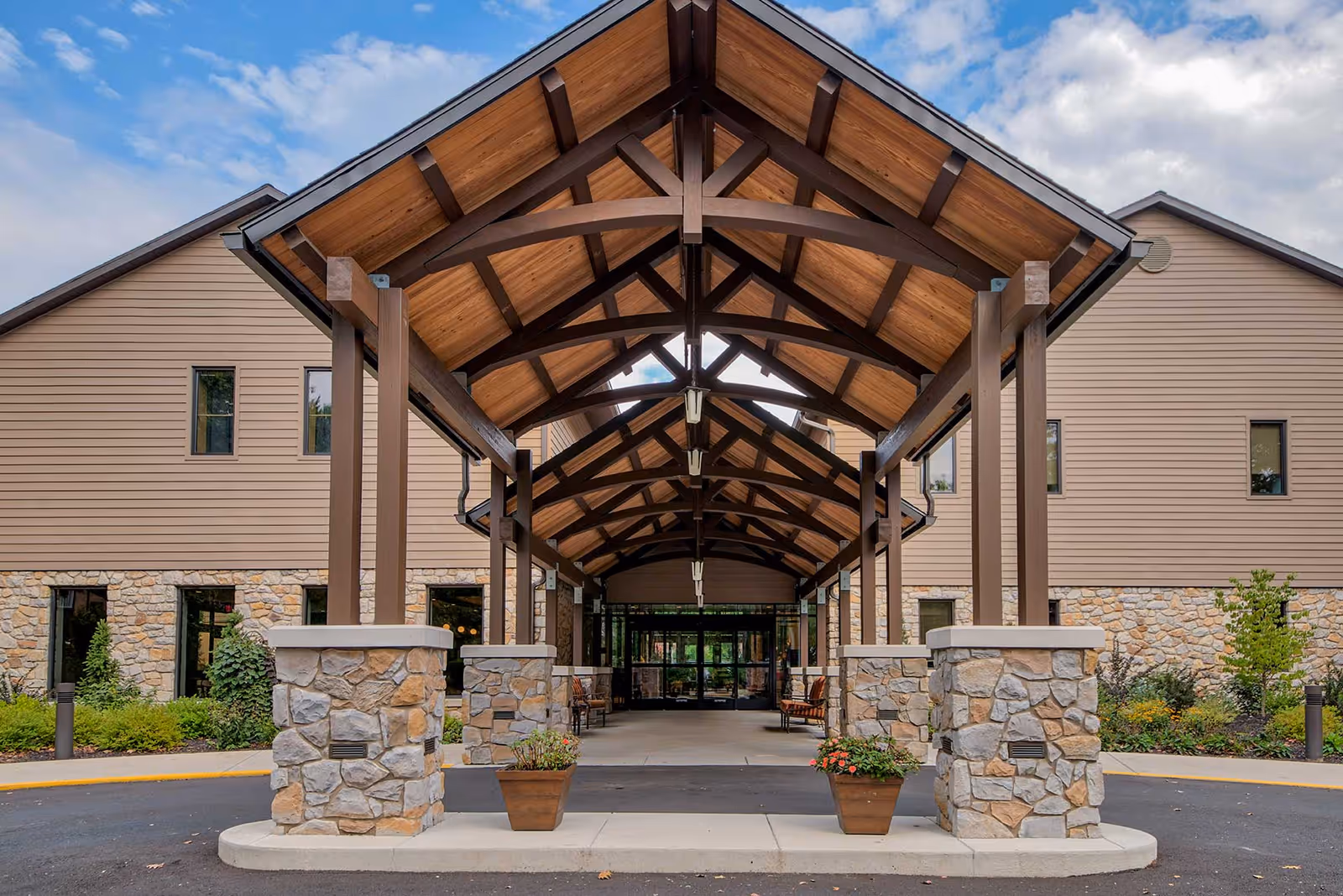 Entrance of a senior living facility with a covered driveway supported by wooden beams and stone pillars, flanked by potted plants and landscaped greenery, under a partly cloudy sky.