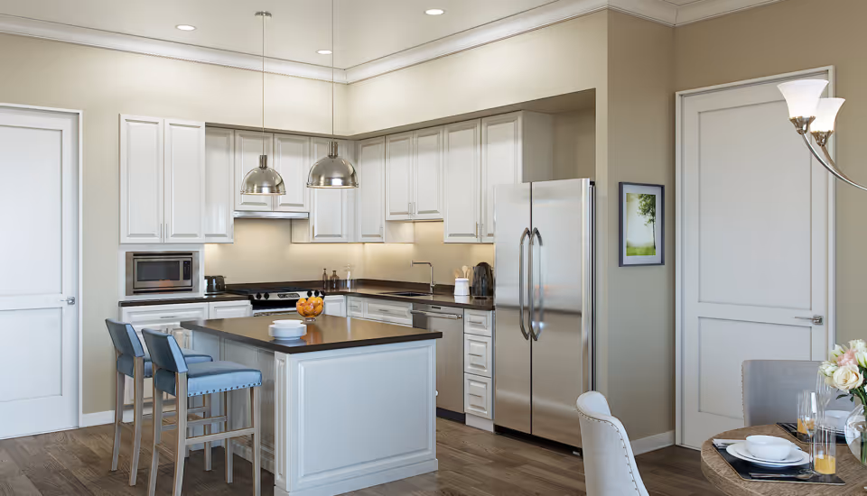 Modern kitchen with white cabinets, stainless steel refrigerator, microwave, and dishwasher. There is a kitchen island with a dark countertop and two blue cushioned bar stools. Two pendant lights hang above the island. Adjacent to the kitchen is a dining area with a round table set with plates, glasses, and a vase of flowers.
