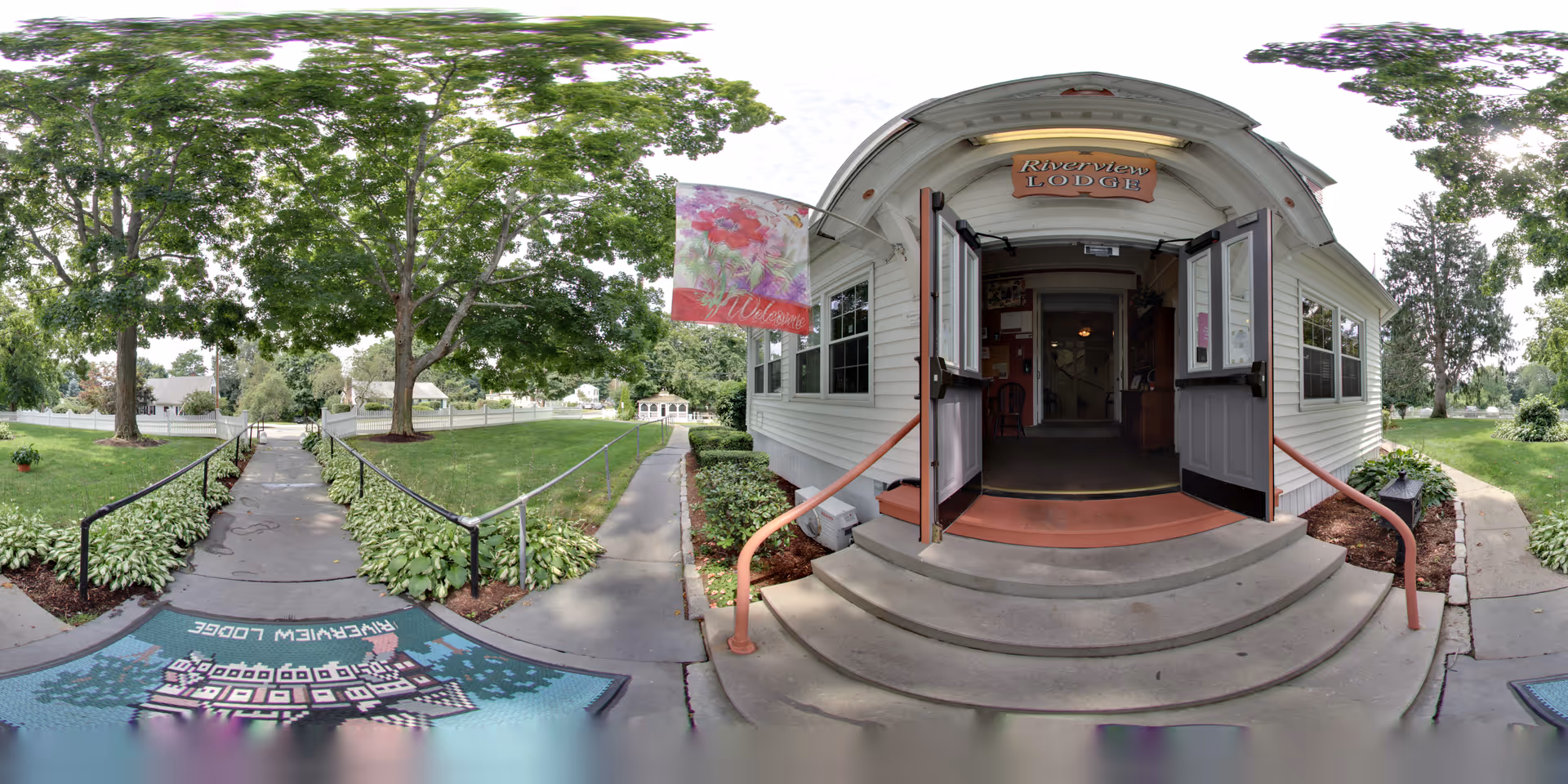 Front entrance of Riverview Lodge Residential Care Home with open double doors, concrete steps leading up to the entrance, a welcome flag with floral design, and a pathway lined with greenery and trees on both sides.
