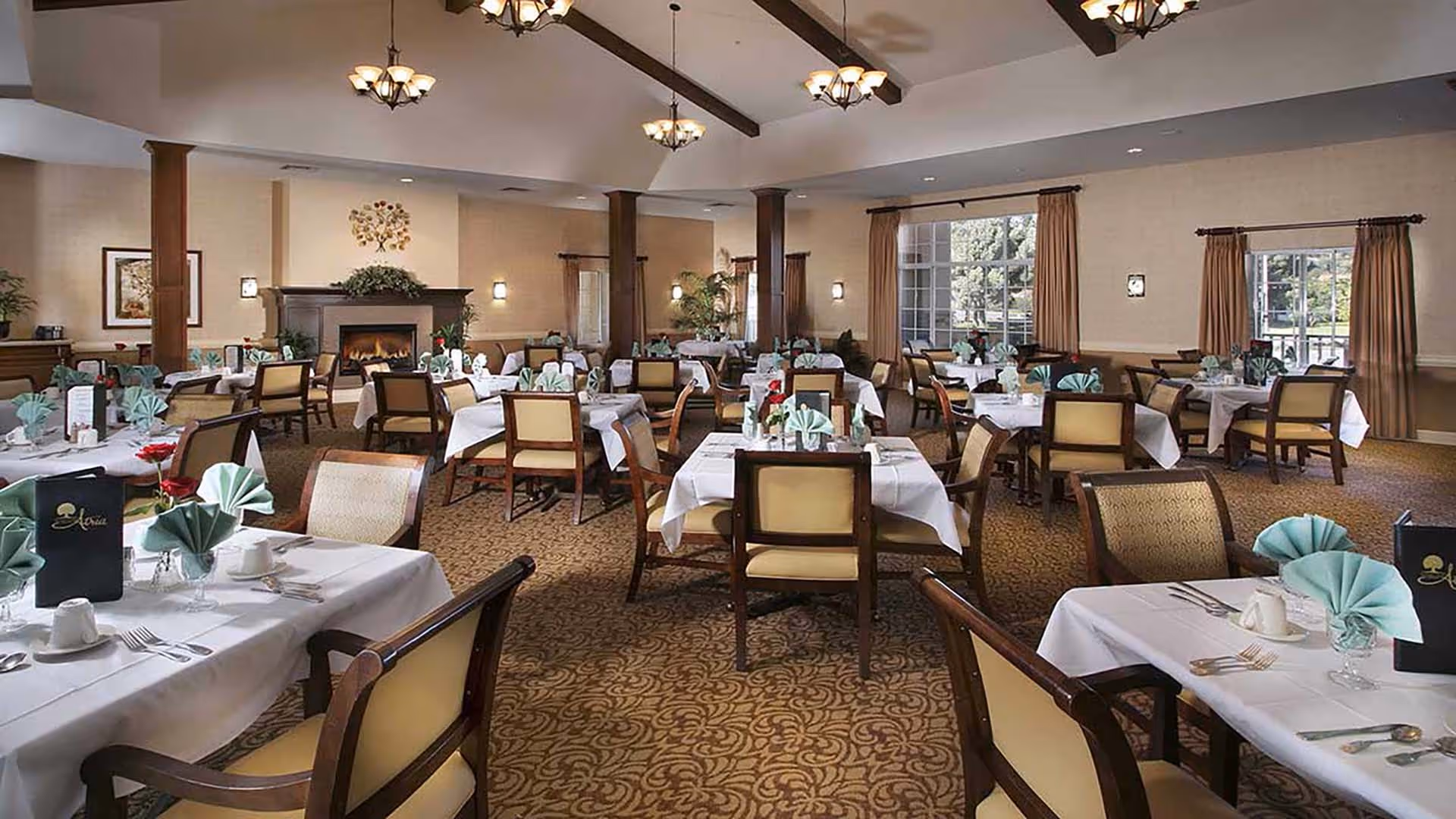 A spacious dining room with multiple tables covered in white tablecloths, each set with folded green napkins, cups, and cutlery. The room features beige walls, large windows with brown curtains, a patterned carpet, wooden chairs with beige cushions, and a fireplace with a decorative piece above it. Ceiling lights hang from exposed wooden beams.
