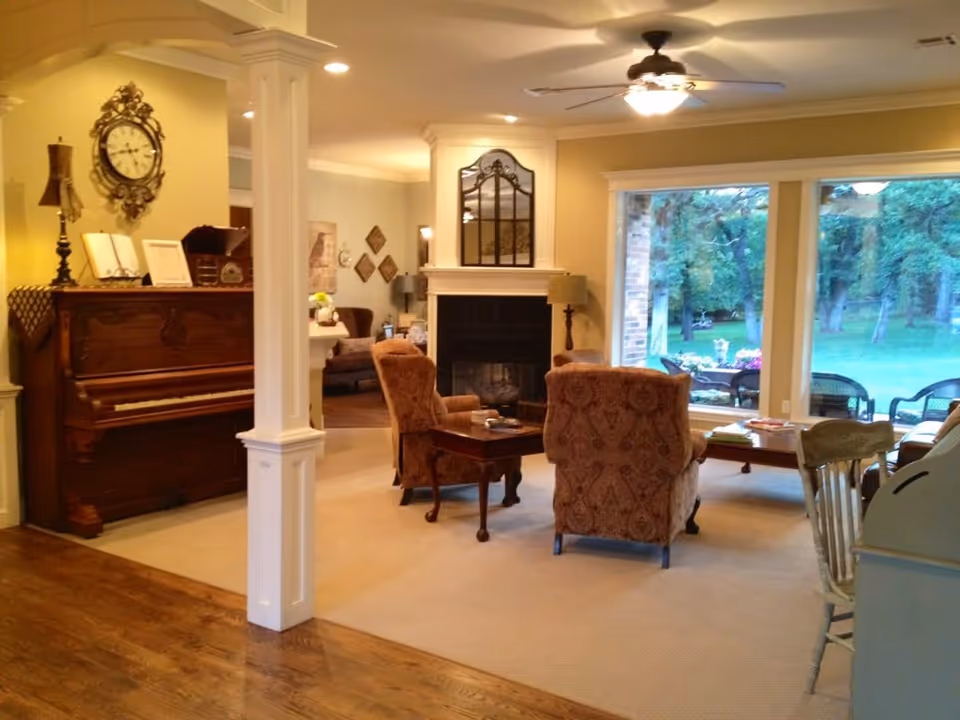 A cozy living room with two patterned armchairs facing a fireplace with a decorative mirror above it. To the left, there is a wooden piano with a vintage clock and a lamp on top. Large windows on the right side provide a view of a green outdoor area with trees and patio furniture. The room has warm lighting and a mix of carpet and hardwood flooring.