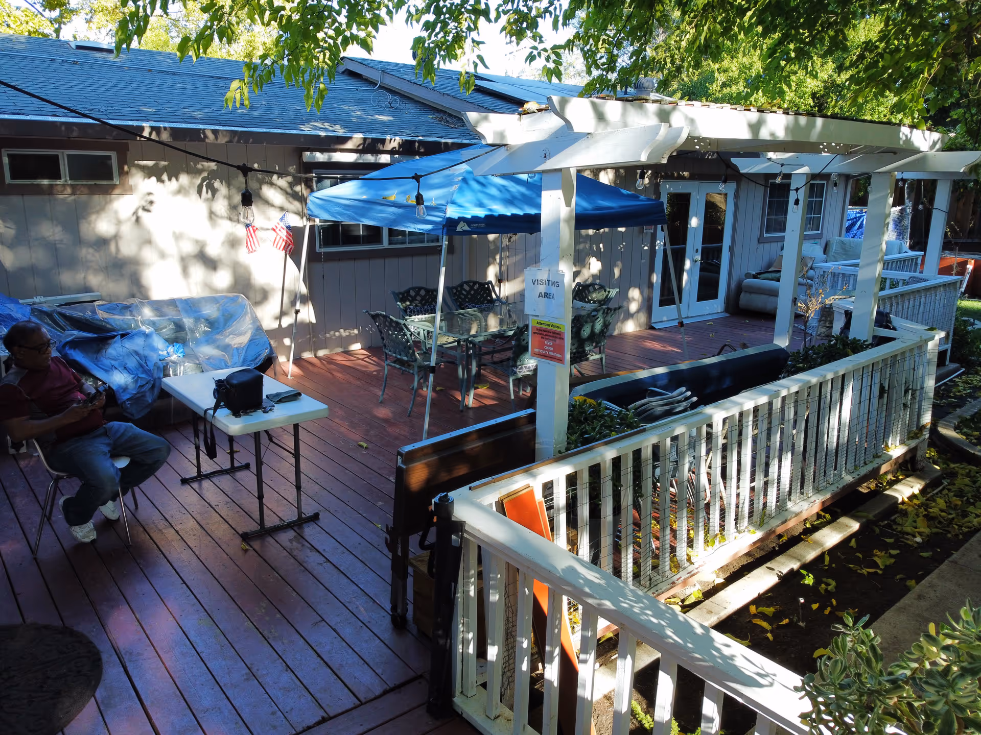 Outdoor patio area with wooden deck, white railing, and a pergola. There is a blue canopy tent covering a glass table with chairs underneath. A man is sitting on a chair next to a white folding table with a black bag on it. The area is shaded by trees and there are string lights hanging from the pergola. A sign on one of the pergola posts reads 'VISITING AREA'.