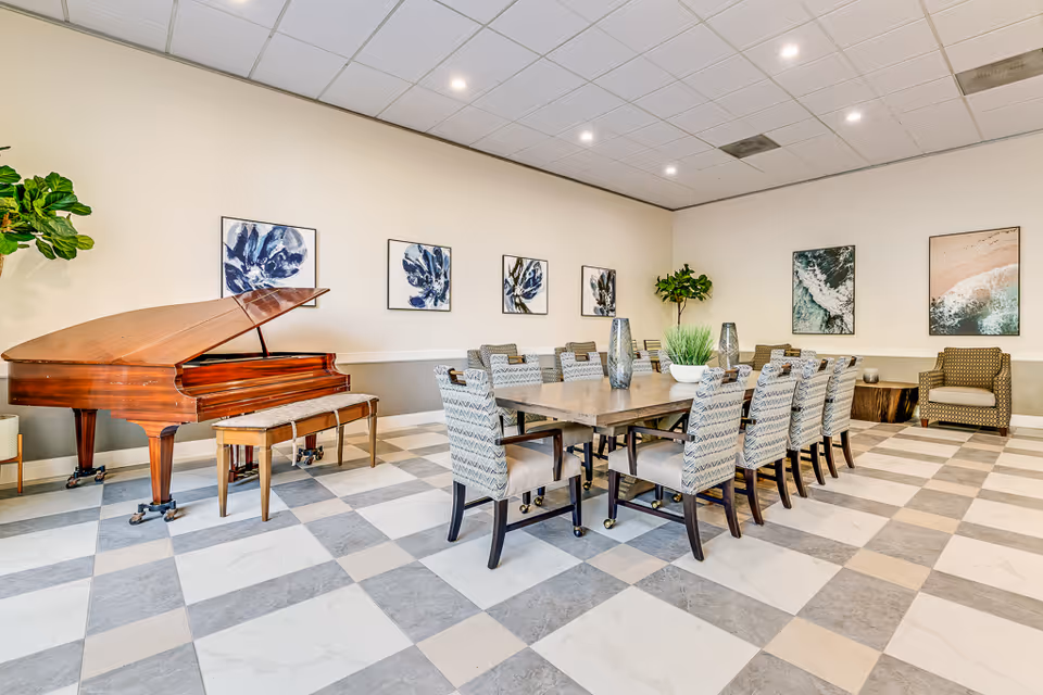 A spacious, well-lit room with a checkered tile floor featuring a large rectangular table surrounded by patterned chairs. A grand piano with a matching bench is positioned to the left, and the walls are decorated with framed artwork. There are also two potted plants and a single armchair in the corner.