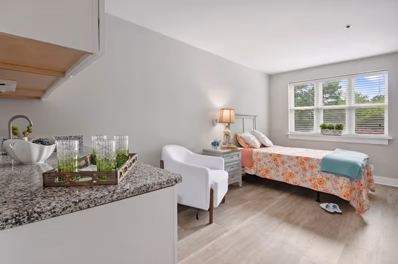 A cozy bedroom featuring a floral bedspread, a bedside lamp, and a window with natural light. A small table with decorative items and a white chair are also visible.