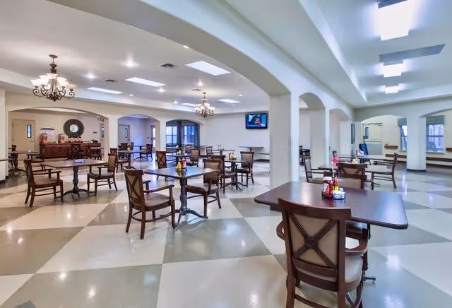 A spacious dining room in a senior living facility with multiple wooden tables and chairs arranged neatly. The room features a checkered tile floor, white walls, and ceiling lights. There are chandeliers hanging from the ceiling and a flat-screen TV mounted on the wall. The room has large windows allowing natural light to enter.