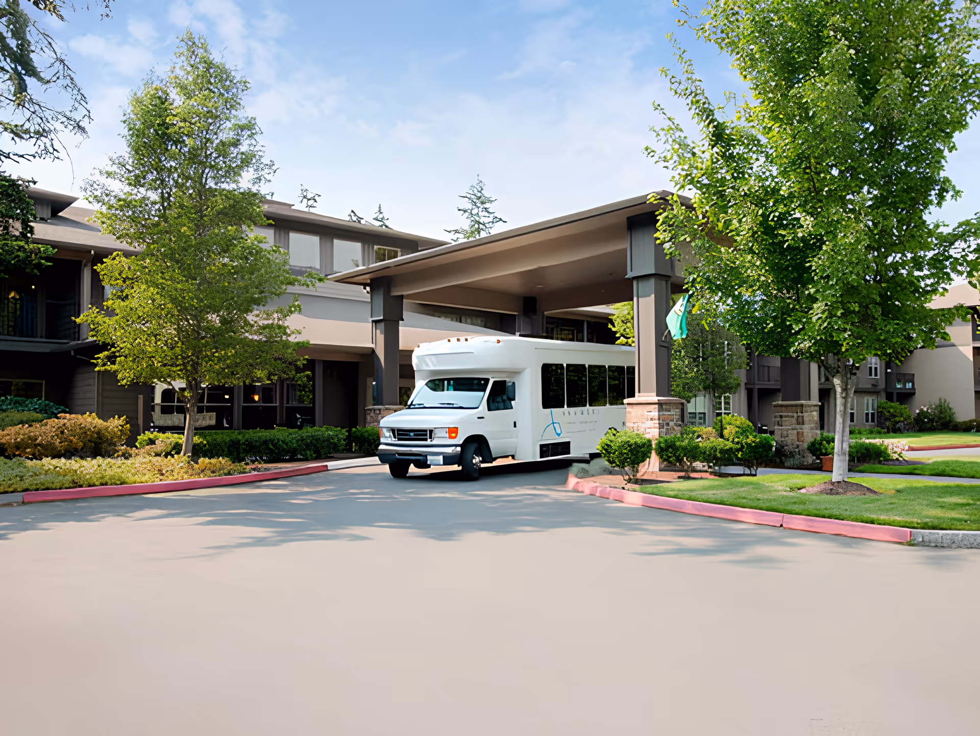 Exterior view of Port Townsend Senior Living by Cogir showing a covered entrance with a white shuttle bus parked underneath. The building is surrounded by green trees and well-maintained landscaping under a partly cloudy sky.