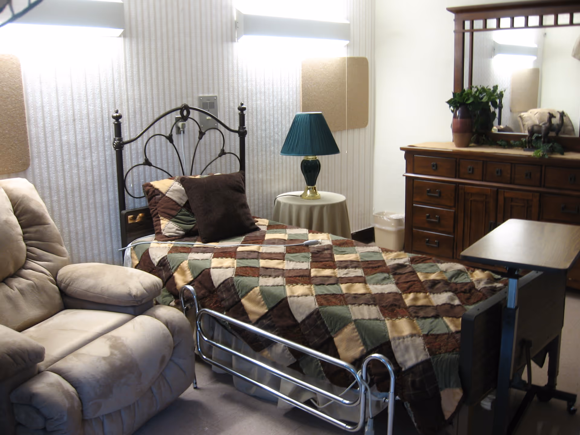 A cozy assisted-living bedroom with a single bed covered by a patchwork quilt, a recliner, a small table lamp, and a wooden dresser with mirror.