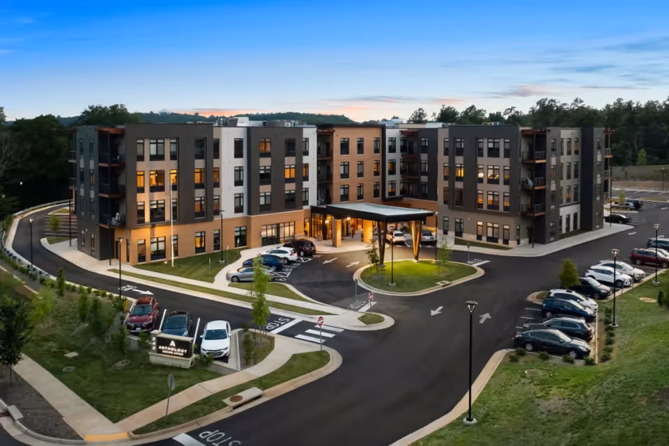 A modern four-story senior living facility building with a covered entrance and several parked cars in the parking lot, surrounded by landscaped greenery and trees under a clear evening sky.