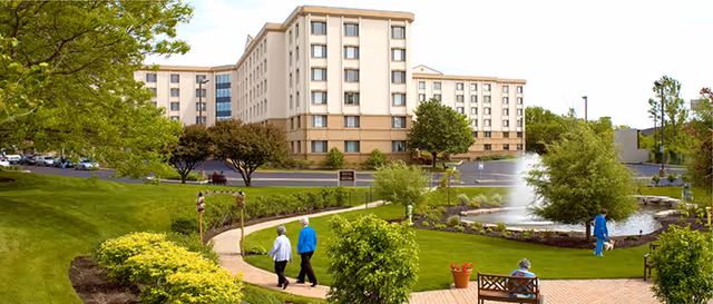 Front view of a multi-story retirement community building with landscaped grounds, a pond and fountain, walkways and people.