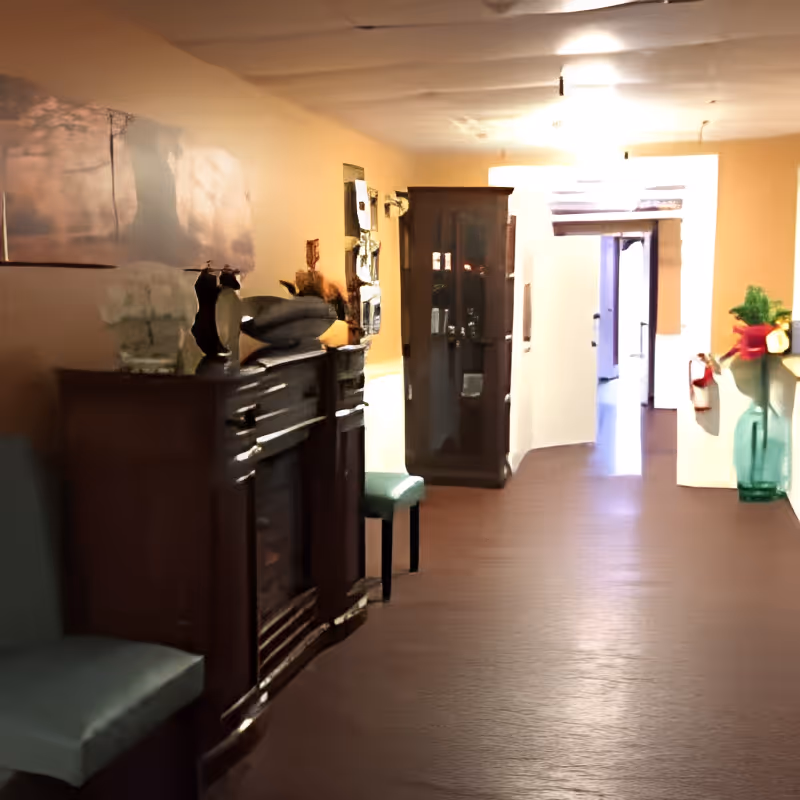 A hallway in a senior living facility with wooden flooring and beige walls. On the left side, there is a dark wooden cabinet with decorative items on top and a green cushioned chair. Further down the hallway, there is a glass-fronted wooden cabinet. On the right side, there is a large green vase with a red ribbon and some greenery. The hallway leads to a bright doorway at the end.