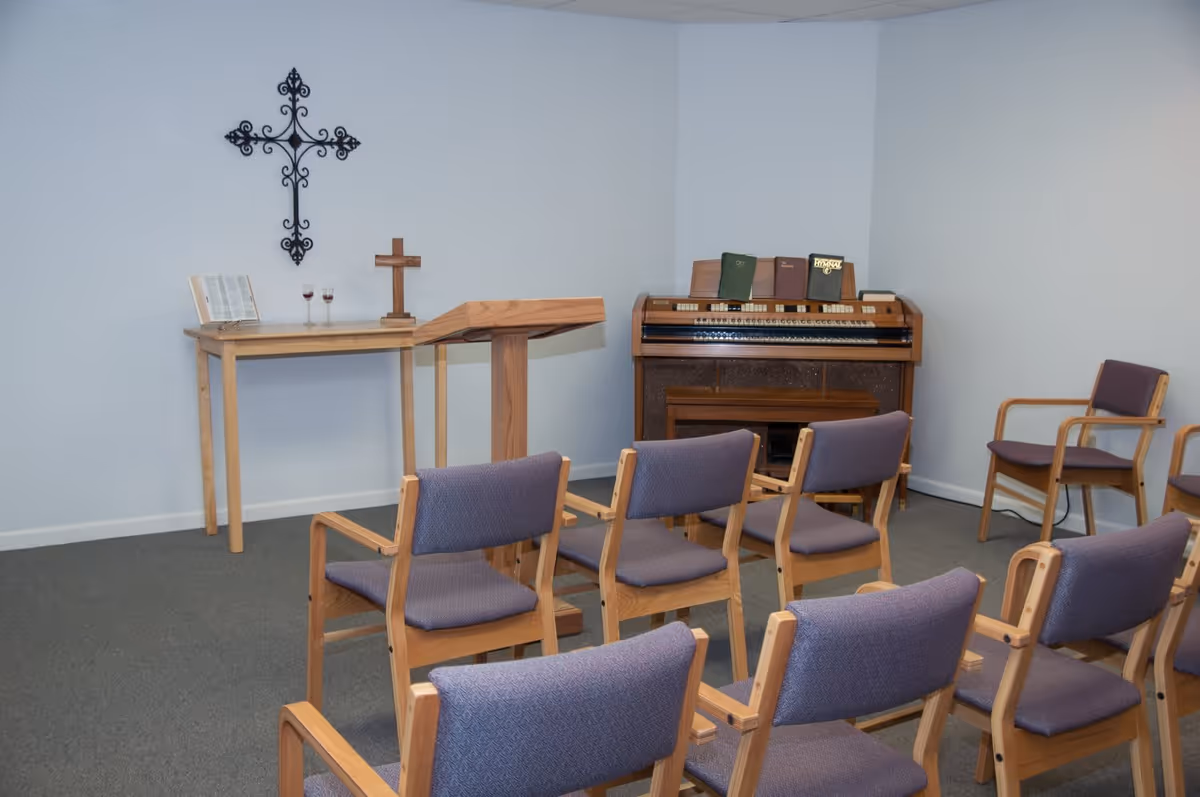 Small chapel room with wooden chairs arranged in rows facing a wooden podium and table. On the table are two wine glasses, an open book, and a wooden cross. A decorative metal cross hangs on the wall above the table. To the right is an organ with hymnals placed on top.
