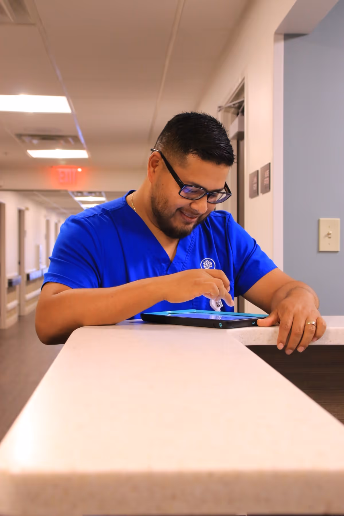 A man wearing blue scrubs and glasses is standing behind a reception desk in a hallway, smiling and interacting with a tablet device.