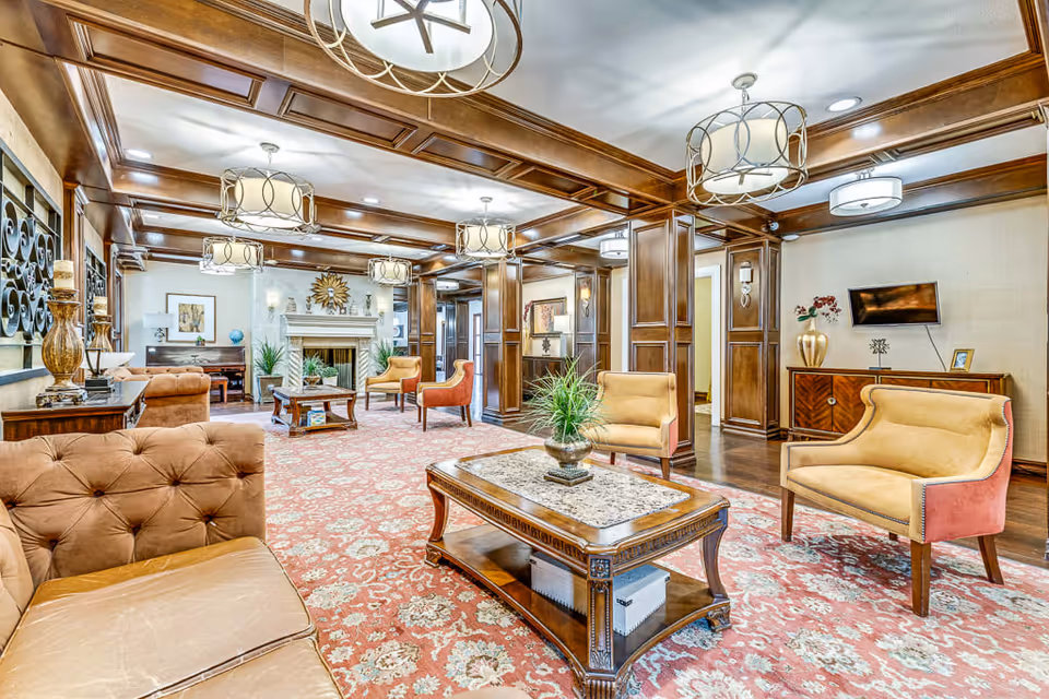 A spacious and elegant senior living facility common area with wooden paneling on the walls and ceiling beams. The room features multiple comfortable upholstered chairs and sofas arranged around wooden coffee tables with decorative plants. A large patterned carpet covers the floor, and several modern light fixtures hang from the ceiling. In the background, there is a fireplace with decorative items on the mantel and framed artwork on the walls.