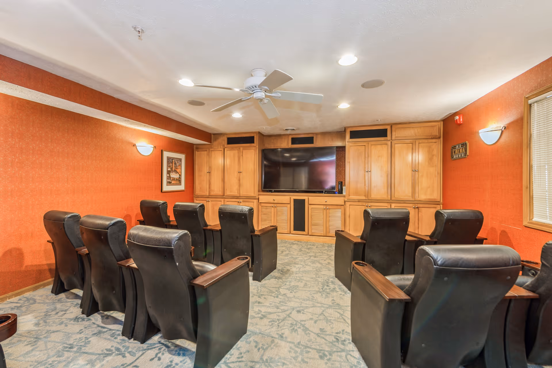 A cozy media room with two rows of black leather recliner chairs facing a large flat-screen TV mounted on a wooden cabinet. The walls are painted a warm orange color, and the ceiling has recessed lighting and a white ceiling fan. There is a framed picture on one wall and a window with blinds on the other.
